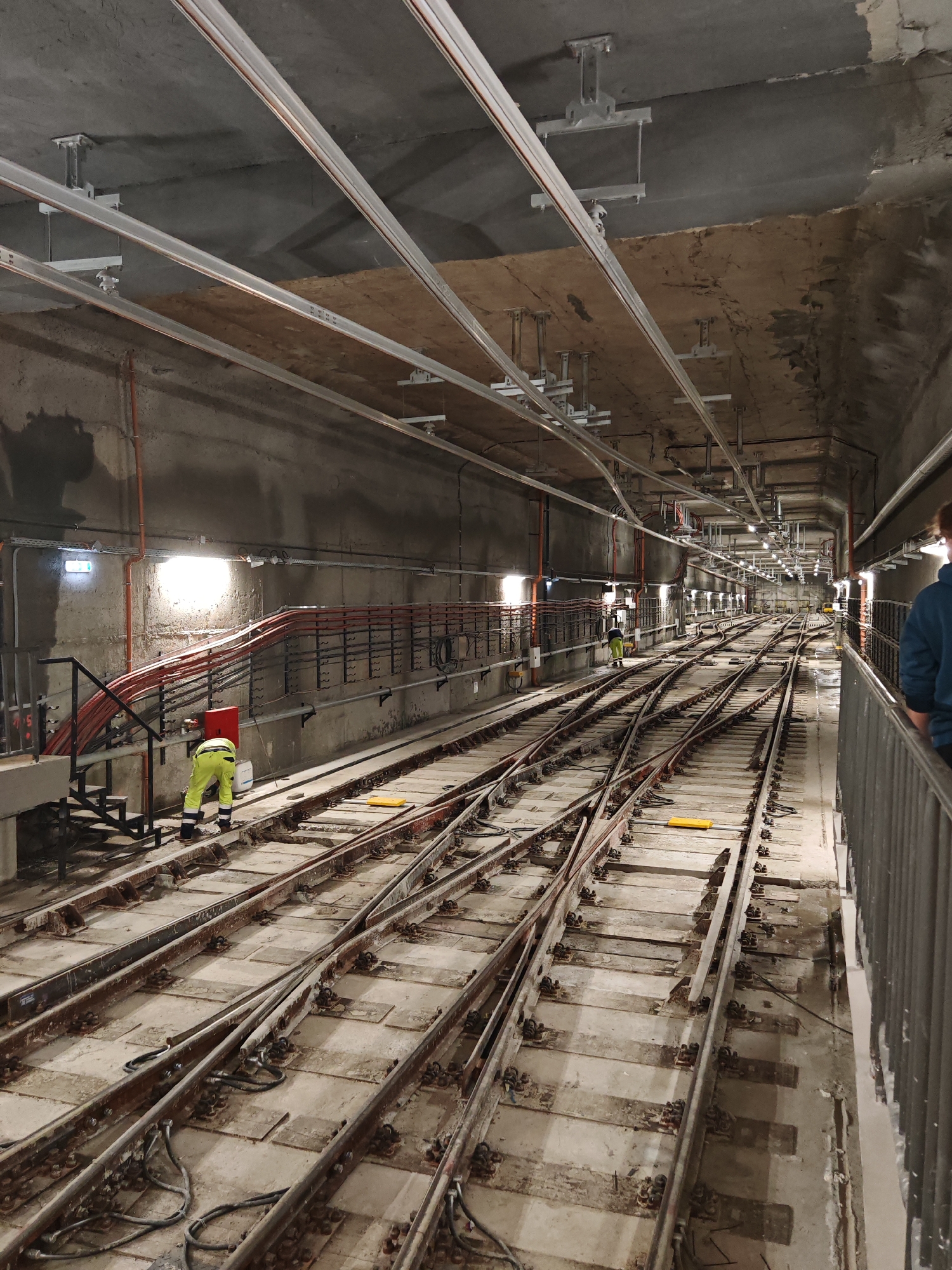 The image depicts a maintenance area within a subway or rail tunnel. Two workers are visible, one in the left foreground wearing high-visibility yellow work clothing and bending over the tracks, and another further down the tunnel, also wearing high-visibility clothing. The tunnel is lined with numerous tracks that converge and diverge. The ceiling is unfinished concrete with support structures for lighting and utilities, including conduits and cable trays running along the walls and ceiling. Orange and red wiring are visible along the left wall. The lighting fixtures are mounted at intervals along the tunnel ceiling. A metal railing is partially visible on the right side of the image. The location appears to be underground due to the tunnel structure and absence of natural light. The location is not clearly discernible.