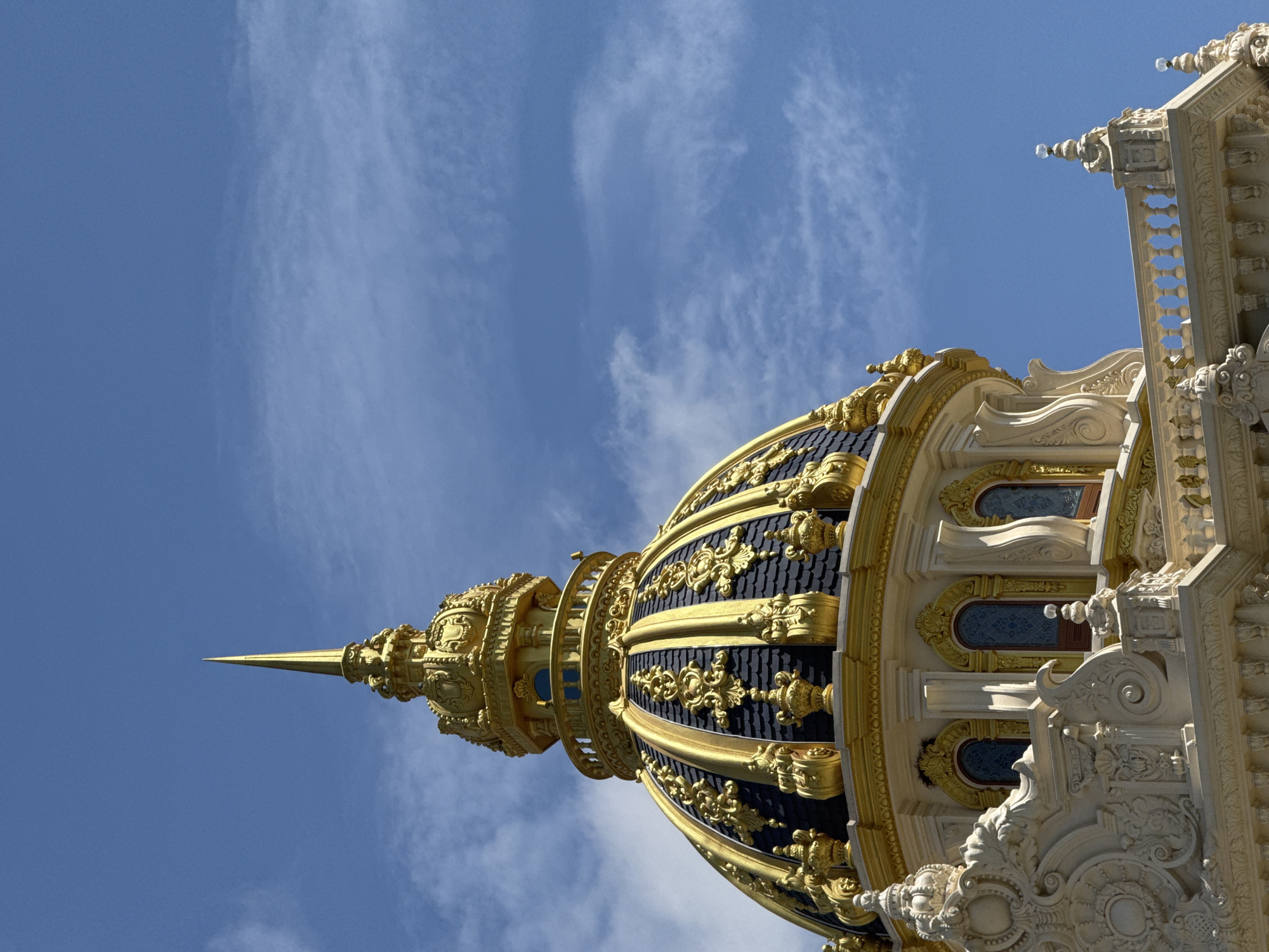The image shows a close-up, low-angle view of the golden dome of the Linh Phuoc Pagoda in Da Lat, Vietnam. The dome is intricately decorated with ornate golden patterns against a dark, tiled background. It is topped with a golden spire. Below the dome, the white facade of the pagoda is visible, featuring elaborate carvings, architectural details, and arched windows with blue glass. A decorative balustrade runs along the edge of the structure. The sky above is a clear blue with scattered white clouds. The scene is brightly lit by sunlight.