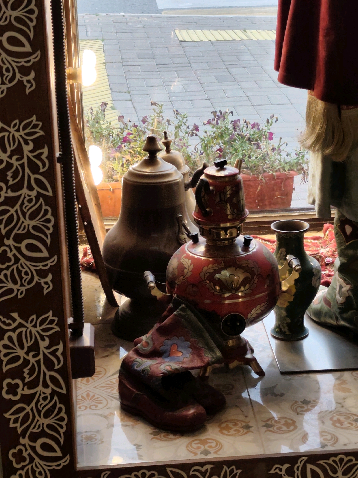 The image captures a close-up view of a display in what appears to be a shop window in Kazan, Russia, showcasing traditional decorative items. In the foreground, a red and gold samovar, a traditional Russian tea urn, is prominently displayed, adorned with intricate floral patterns. Next to it is a green vase with similar decorative motifs. To the left of the samovar, a large, dark bronze coffee pot or jug stands, also featuring ornate detailing. In front of the samovar, a pair of burgundy leather shoes are placed, with a patterned cloth draped over them. The items are set against a tiled floor with a decorative pattern.
Through the window, a glimpse of the outside world reveals a paved area with grey and yellow tiles, suggesting a pedestrian walkway or street. Flower pots filled with purple flowers are visible outside, adding a touch of color to the background. A red fabric, possibly a curtain or part of a garment, hangs from the top right of the frame, obscuring a portion of the background and a figure standing there. The lighting suggests it is daytime, with natural light filtering through the window and artificial lights from within the shop contributing to the illumination. The overall scene evokes a sense of cultural heritage and artisanal craftsmanship, likely presented for sale.