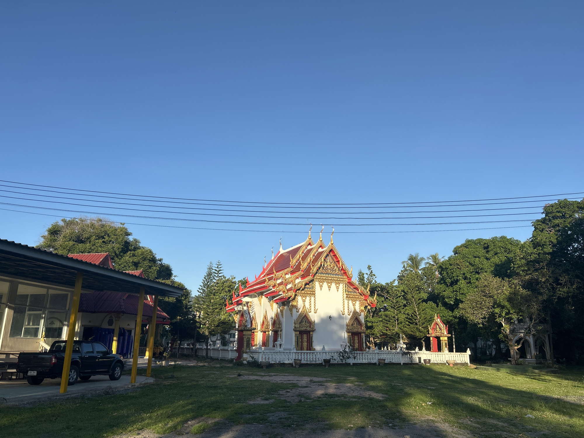 The image depicts a Buddhist temple in Tambon Ko Pha Ngan, Thailand, under a clear blue sky. The temple features a white structure with ornate gold and red roofing, flanked by a smaller red and gold shrine. In the foreground, to the left, is a building with a red roof supported by yellow posts, under which a black pickup truck is parked. The area is surrounded by lush green trees and a grassy lawn. Power lines are visible stretching across the upper portion of the frame.