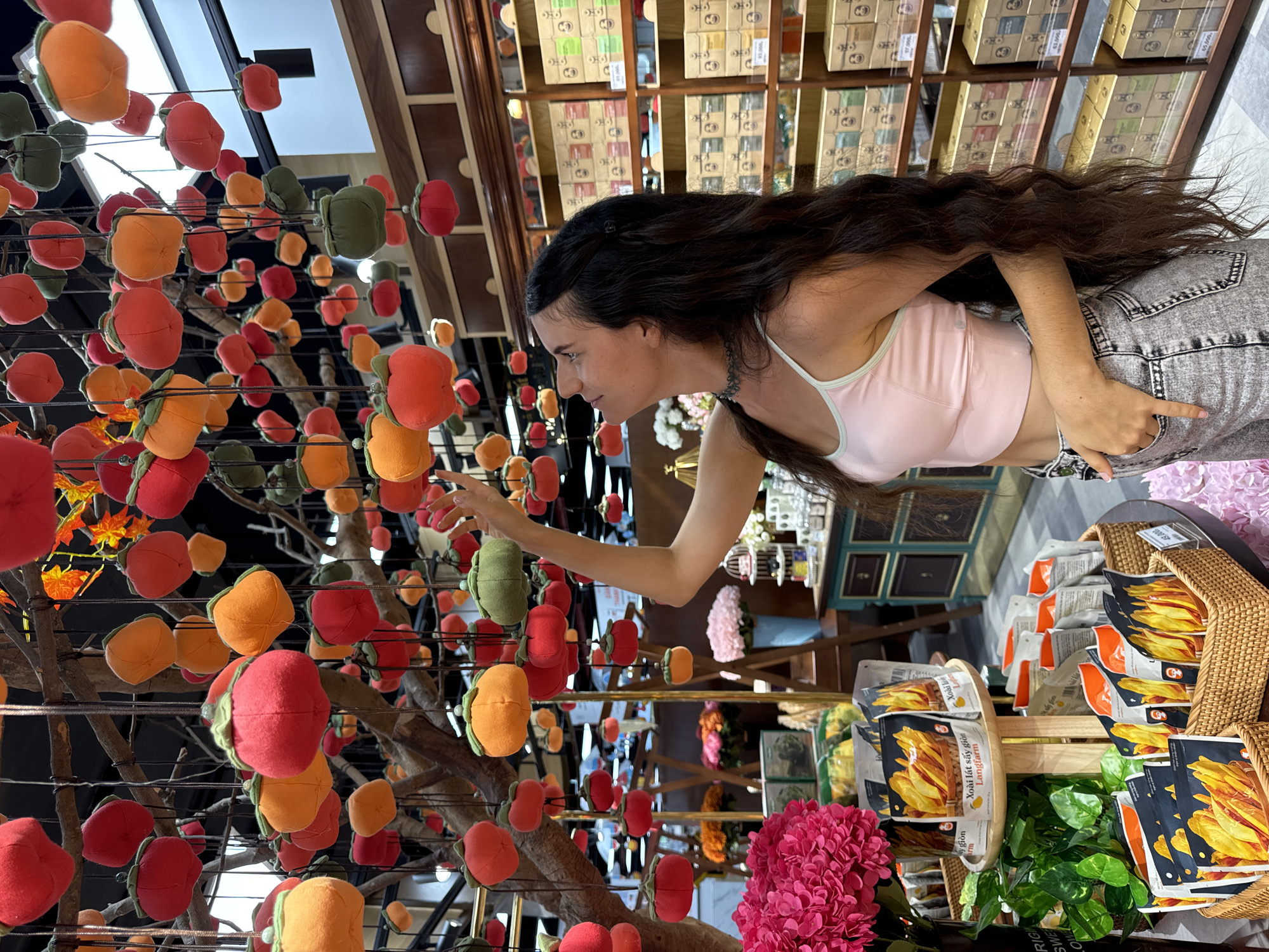 A woman with long, dark hair reaches toward a tree adorned with fabric fruit ornaments in what appears to be a retail setting in Phu Thuy Ward, Vietnam. She is dressed in a light pink tank top and grey jeans. The "tree" is centrally positioned in the shot, acting as a focal point. The interior space is filled with various merchandise, including packaged food products in baskets and neatly arranged boxes on shelves. A pink artificial flower arrangement is visible near the bottom of the frame.