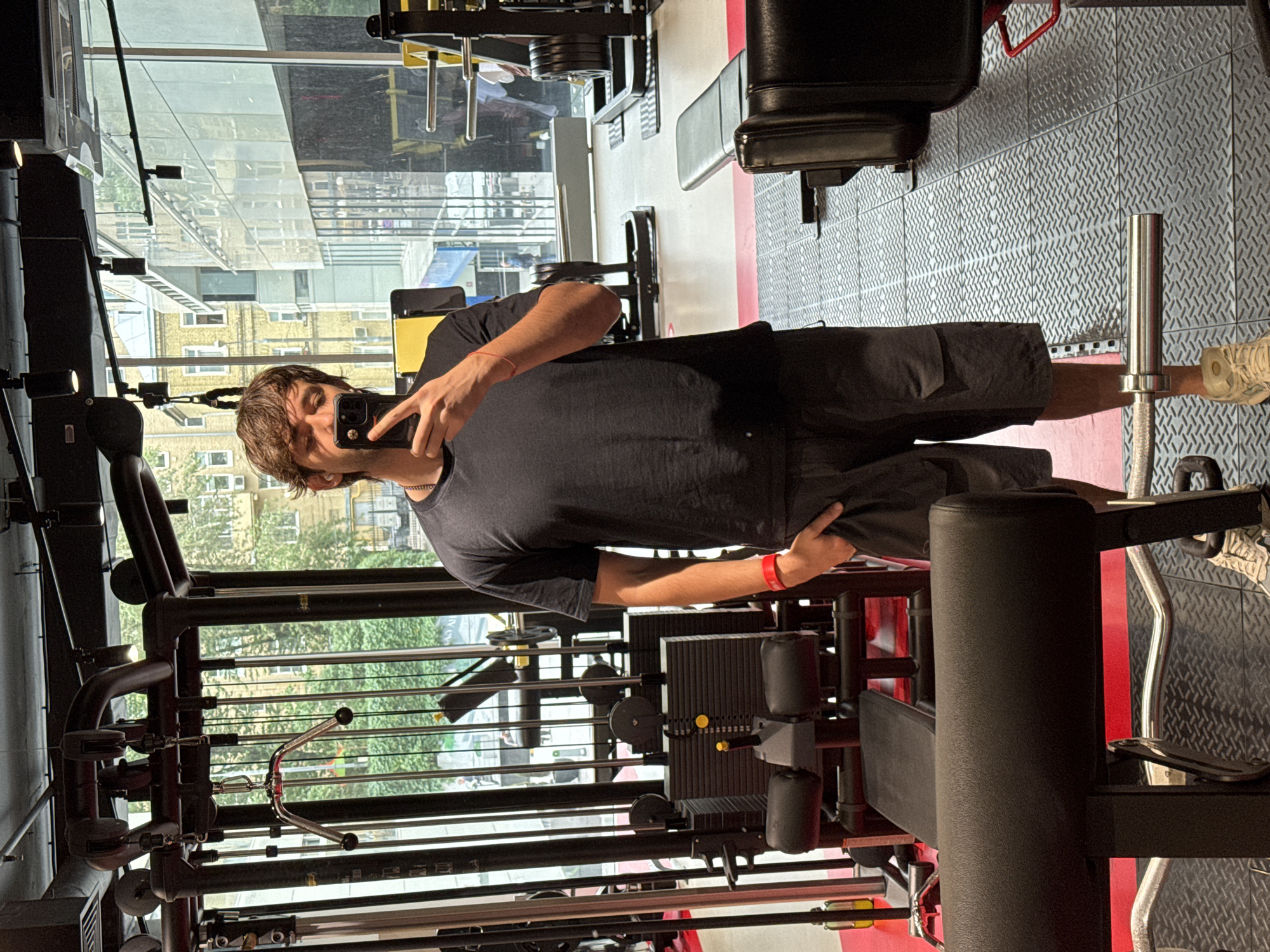 A young man is taking a selfie in a gym. He is wearing a black t-shirt and shorts. The gym appears to be well-equipped with various weight machines and benches. Through the large windows behind him, the exterior of a building and trees are visible, suggesting it is daytime. The man is holding a smartphone in his right hand, capturing his reflection in a mirror or a reflective surface of the gym equipment. He has dark, messy hair and is looking towards the camera with a neutral expression. A red wristband is visible on his left wrist. The gym floor is covered with a textured matting. The overall scene suggests a casual workout session in an urban environment. The location context of Moscow, Russia is provided, but there are no specific visual cues within the image that confirm this.