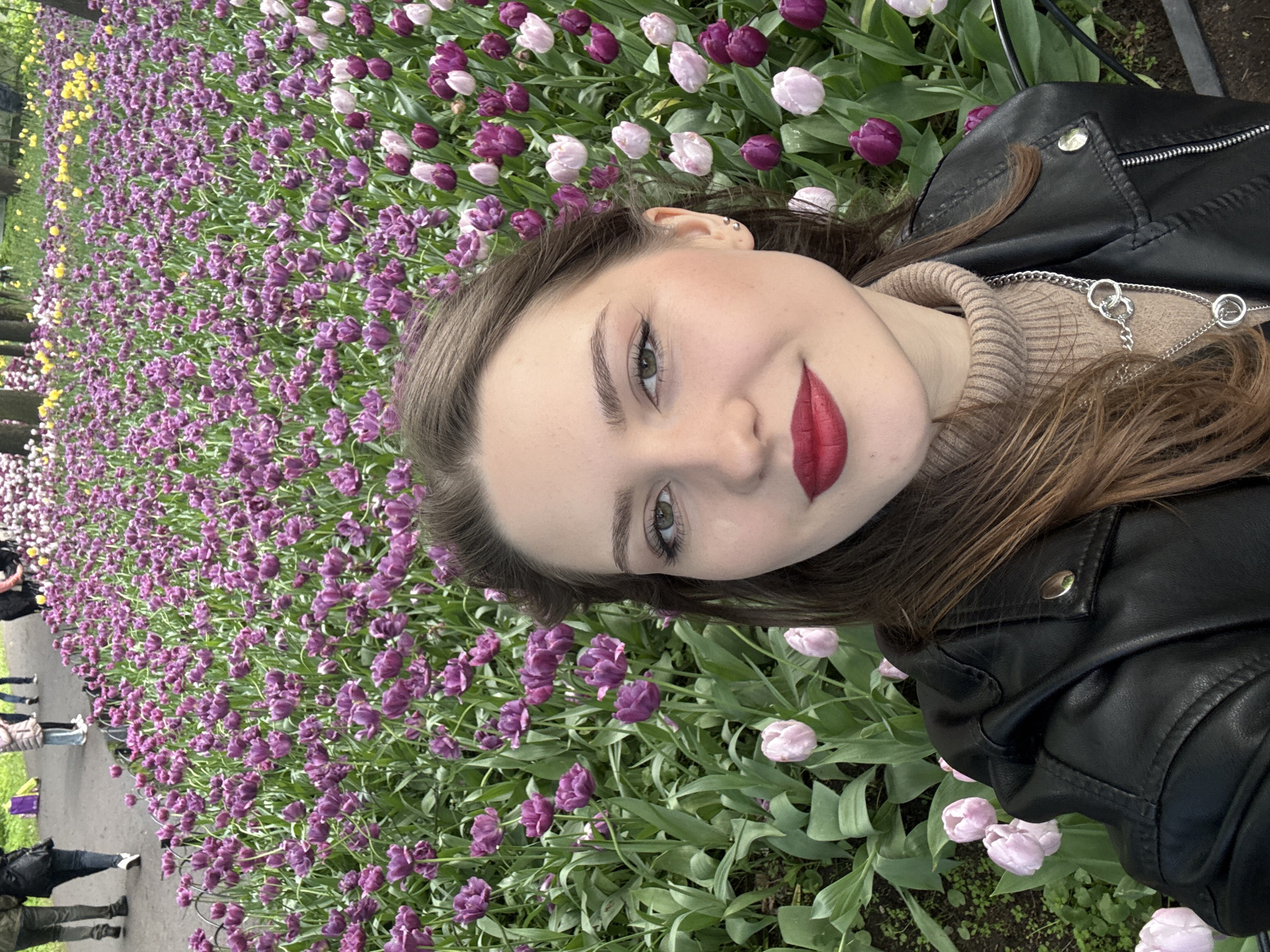 A woman with long brown hair, red lipstick, and a black leather jacket poses for a selfie in a field of purple and pink tulips. The tulips appear to be planted in a garden area. In the background, a group of people are walking along a paved path near more yellow tulips. Based on the tulips and the architecture in the background, the location may be St. Petersburg, Russia, though this cannot be definitively confirmed.