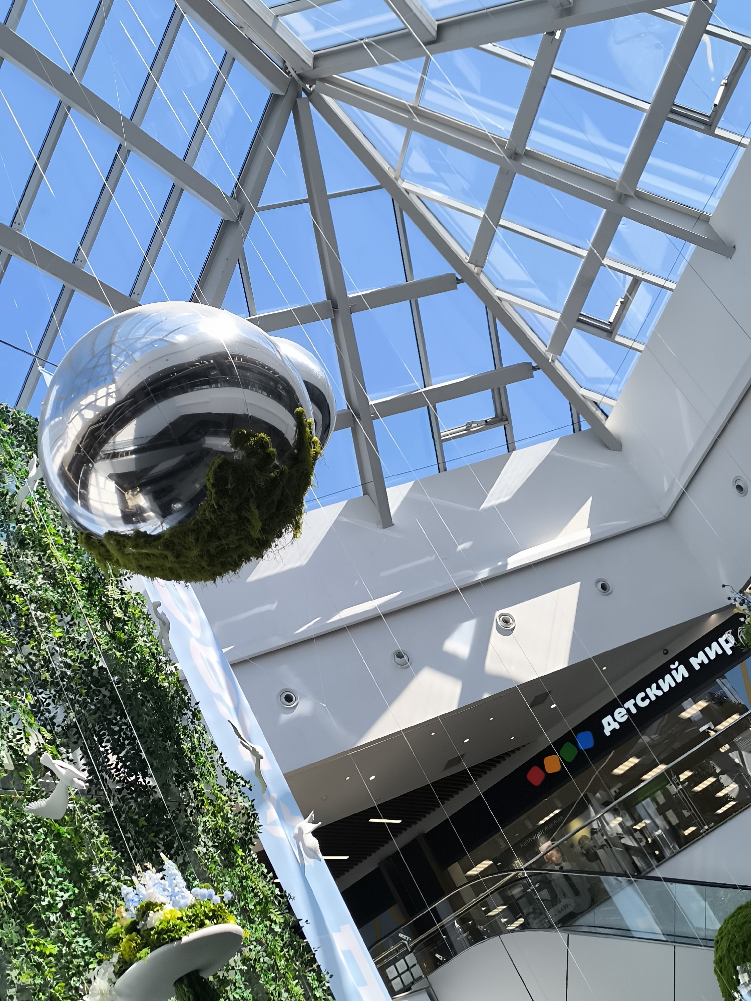 The image is a low-angle, interior shot of a modern shopping mall, likely in Qarağandı city administration, Kazakhstan. A large, mirrored sphere, partially covered in green moss, is suspended from the ceiling by thin wires. It reflects the structure of the mall's glass and metal roof, which allows natural light to filter through. To the left of the sphere, a vertical installation of lush green foliage is visible, adorned with several white bird figurines. In the background, a store sign displaying "детский мир" (Children's World) is illuminated, with a colorful logo consisting of red, yellow, green, and blue squares. Escalators and interior mall architecture are also visible. The overall impression is one of a bright, spacious, and contemporary commercial environment.
