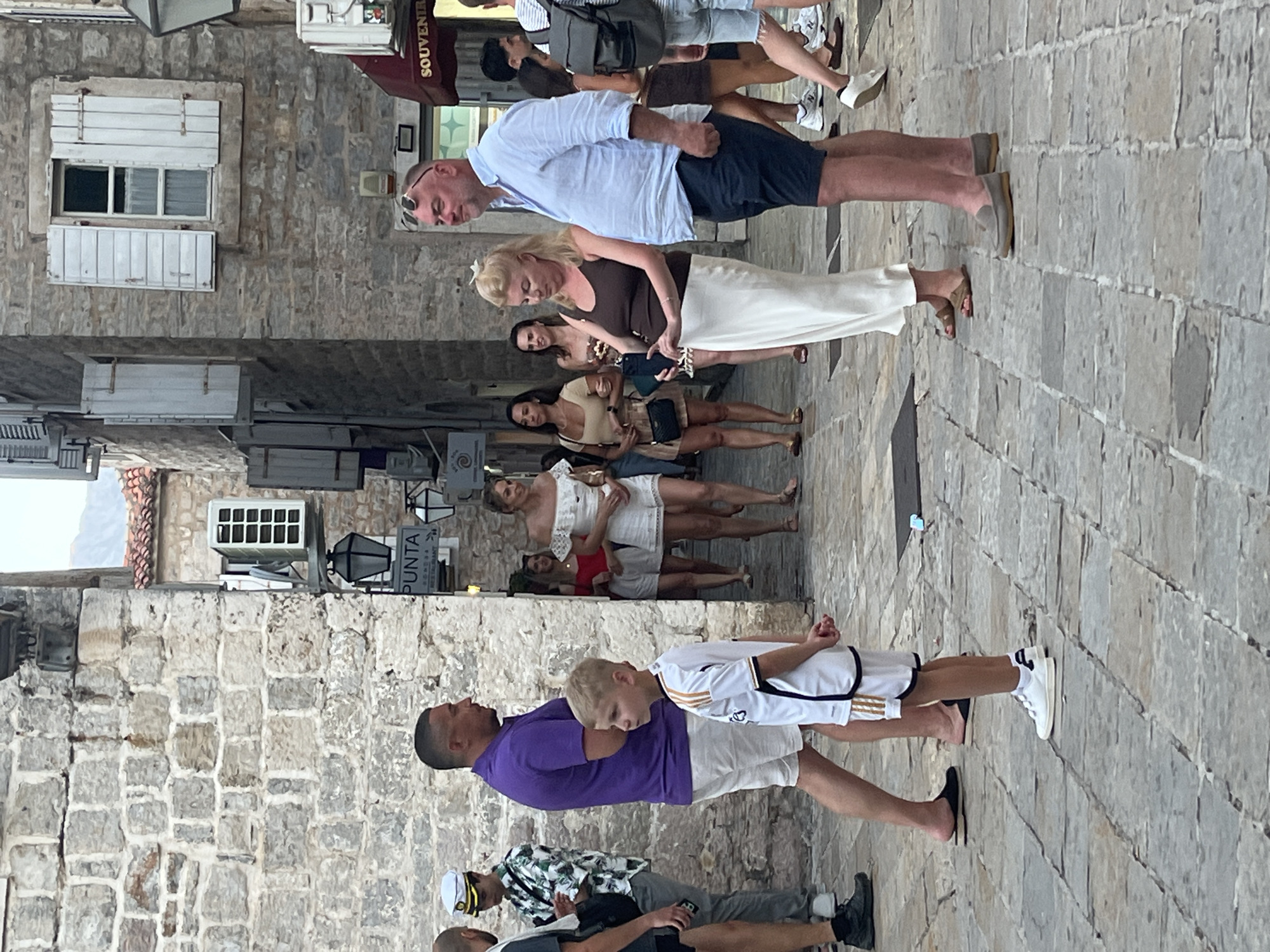 The image depicts a street scene in the old town of Budva, Montenegro, characterized by stone buildings and cobblestone pavement. In the foreground, a young boy wearing a white and gold soccer jersey and shorts stands facing left. To his left, a man in a purple polo shirt and white shorts walks away from the camera. Further into the scene, a group of people are walking. On the right, a man in a light blue button-down shirt and dark shorts stands with a woman in a brown tank top and a long white skirt. Behind them, other individuals are visible walking in the narrow street. The architecture suggests an old European town, with stone walls, wooden shutters on windows, and outdoor lighting fixtures.