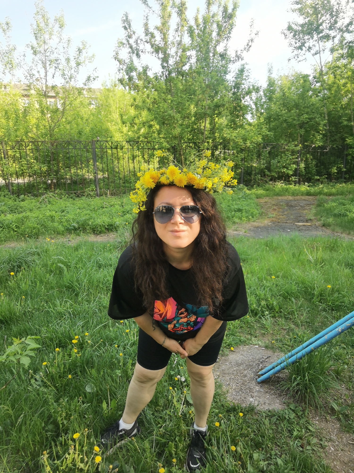 A full shot depicts a woman with long, dark curly hair wearing a crown of yellow dandelions and sunglasses, standing in a grassy area. She is wearing a black t-shirt with a colorful design and black athletic shorts. Her hands are clasped in front of her. She is wearing black athletic shoes with white socks.
The location appears to be a grassy yard with dandelions. In the background, there is a metal fence and lush green trees. A building is visible behind the trees. To the right, there is a piece of blue playground equipment.
Based on the presence of the dandelions, fence, building and playground equipment, the scene is possibly in a residential or park area within the city of Perm, Russia.