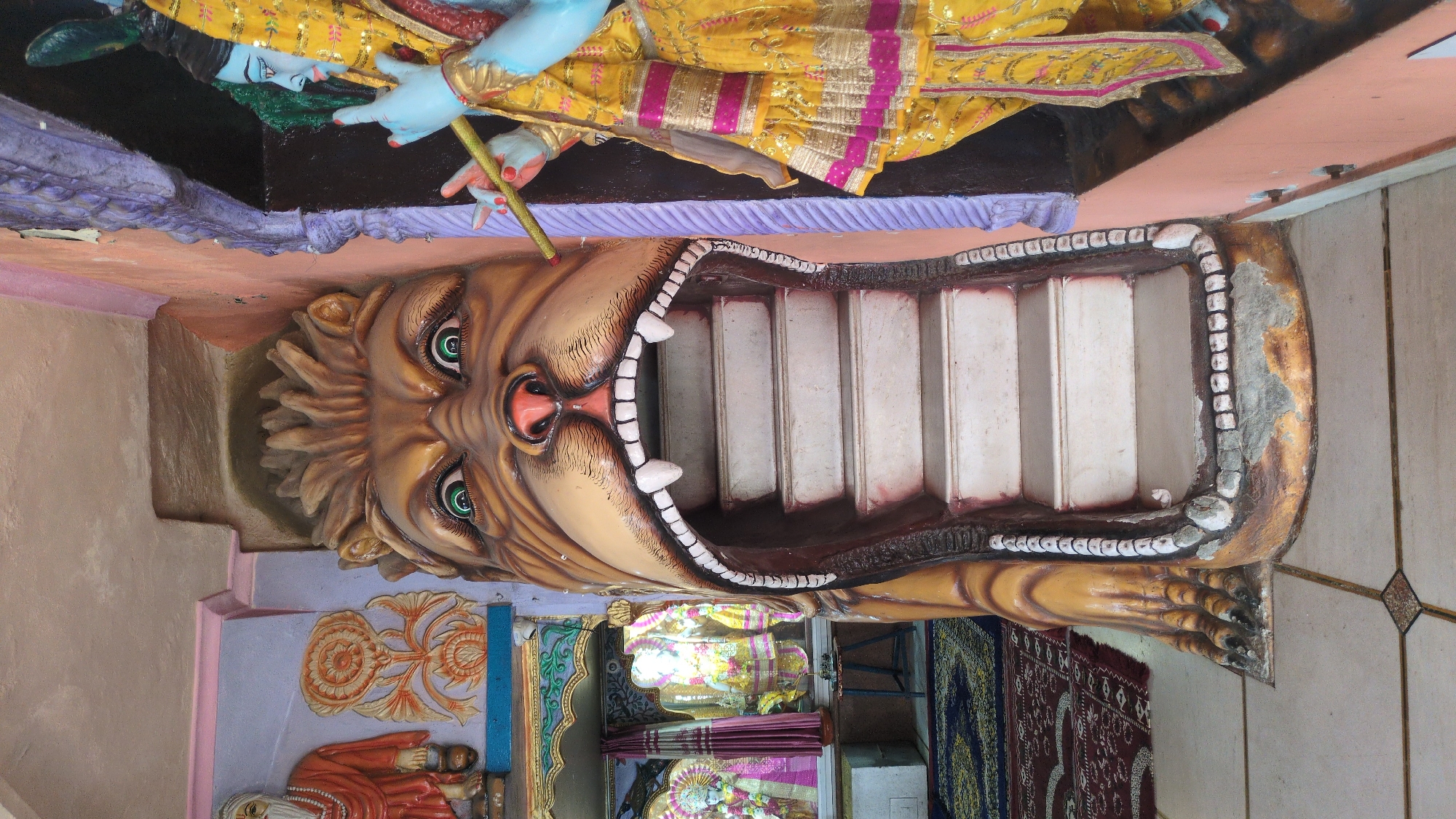 The image captures the vibrant and ornate interior of a Hindu temple, likely located in Bhagsu, India. The central and most striking feature is a large, colorful sculpture of a mythical creature, resembling a lion or Narasimha, with its mouth wide open to form a staircase. The creature's head is intricately detailed with large green eyes, a pink nose, a textured brown mane, and prominent white fangs, while its lower jaw extends into the structure of the stairs. The staircase itself consists of several white steps leading upwards.
Above and to the left of this creature, a statue of a blue-skinned deity, possibly Krishna or Vishnu, is visible. This deity wears elaborate yellow and pink robes with gold detailing and holds a staff. Its face is depicted with a serene expression, and its hands are in a graceful pose. Further to the left, partially visible, is another statue of a figure with an orange robe and a white beard, possibly a sage or Hanuman, set against a wall adorned with intricate patterns.
In the deeper background, more colorful statues of deities and various temple decorations are faintly discernible. The floor is covered with patterned mats in shades of red and blue, indicating a space for worship. The walls are painted in varying hues of peach, purple, and beige, contributing to the overall rich and colorful aesthetic of the temple. The scene is brightly lit, highlighting the detailed artistry of the sculptures and decorations. There is no visible text in the image.
