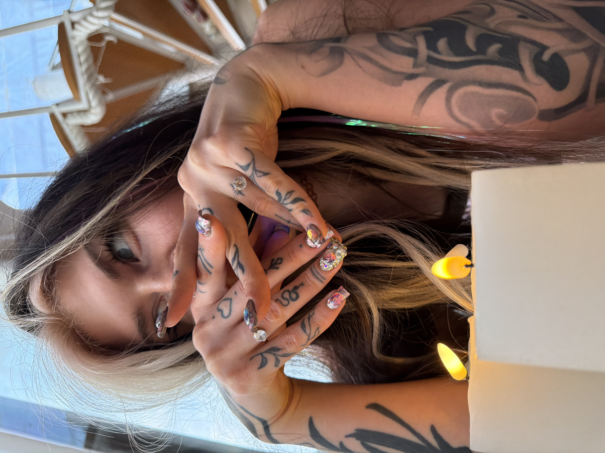 A woman with dark hair and a blonde streak, tattooed arms and hands, and elaborate nail art is centrally positioned in the image. She is holding her hands near her face. A white object with lights is next to her, and a structure with white ropes is in the background, against a blue sky. It is outside at daylight. This is in Budva, Montenegro.