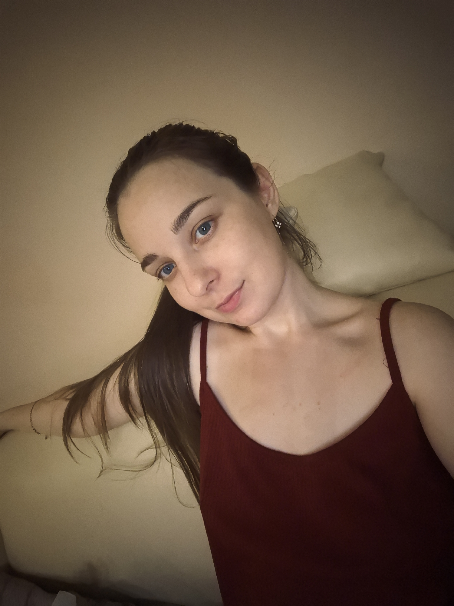 A woman with fair skin, blue eyes, and brown hair pulled back into a ponytail is seen in a selfie. She is wearing a dark red tank top. The background is a cream color, likely a wall, and a pillow is visible behind her head. The lighting is soft, suggesting indoor conditions. There is no indication of the location being in Moscow, Russia, or any other specific city.