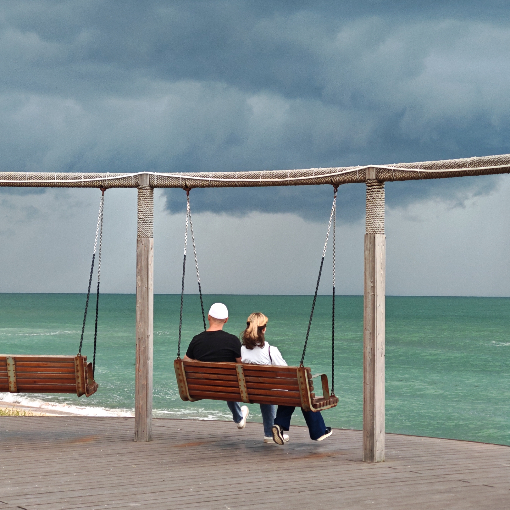 A medium shot depicts a man and woman seated on a wooden swing bench at the edge of a wooden deck platform. Another empty wooden swing is to the left of the couple. The deck overlooks a view of the ocean that has a green tint. The sky is overcast with grey stormy clouds. The scene is on the edge of the ocean at an unknown location.