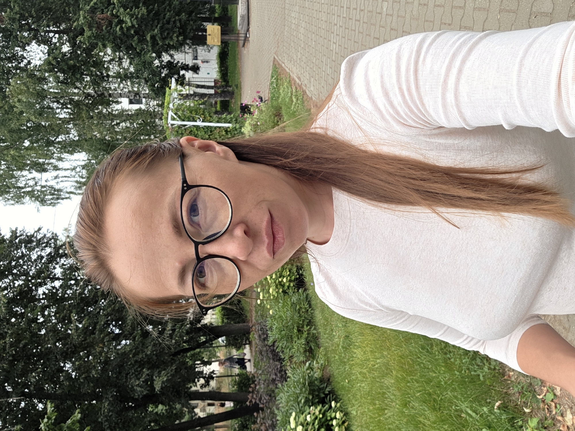 In a close-up, outdoor, selfie-style photograph taken in Chaikovsky, Russia, a woman with long, reddish-brown hair pulled back and wearing black-rimmed glasses is the central subject. She has light skin and is looking towards the camera with a neutral expression. She is wearing a light-colored, long-sleeved, textured shirt. The background features a green lawn, garden beds with lush plants and some white flowers, and trees. A paved walkway with a brick pattern is visible on the right side of the frame. Further in the background, a white structure resembling an archway or pergola can be seen, along with a yellow information sign on a stand and a building. In the middle ground, an older woman in a hat and carrying a bag is walking away from the camera on a path. The overall scene depicts a public park or garden area.
