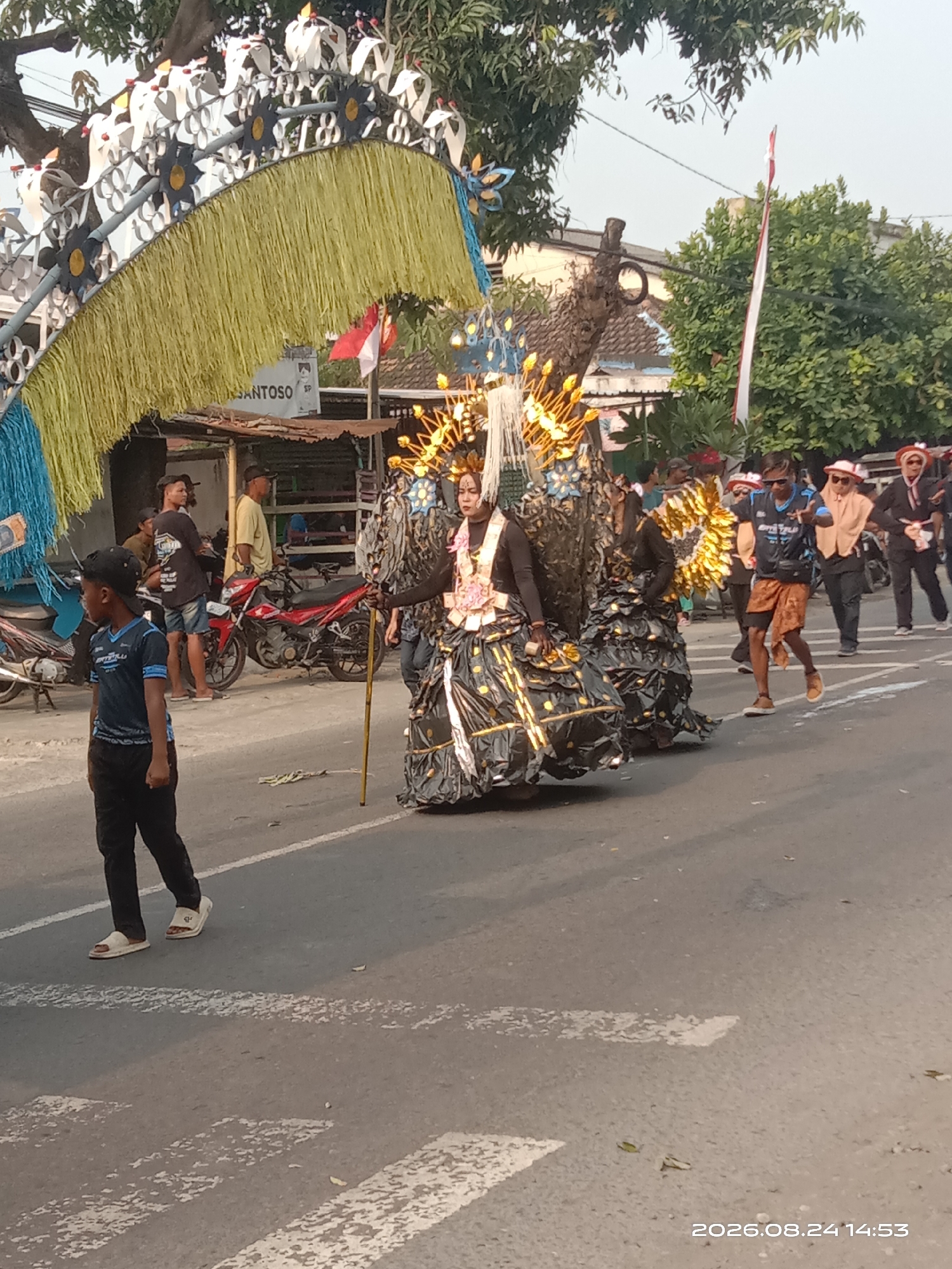 The image shows a street parade in Gudo, Indonesia, on August 24, 2026, at 2:53 PM. A young boy wearing a blue t-shirt and black pants walks across a crosswalk in the foreground. Behind him, a person in a large, elaborate black costume adorned with gold decorations and a crown is being paraded. The costume appears to be made of layered fabric and possibly metallic materials. In the background, there are other parade participants, onlookers, parked motorcycles, and small shops lining the street. The event is taking place outdoors during daylight.