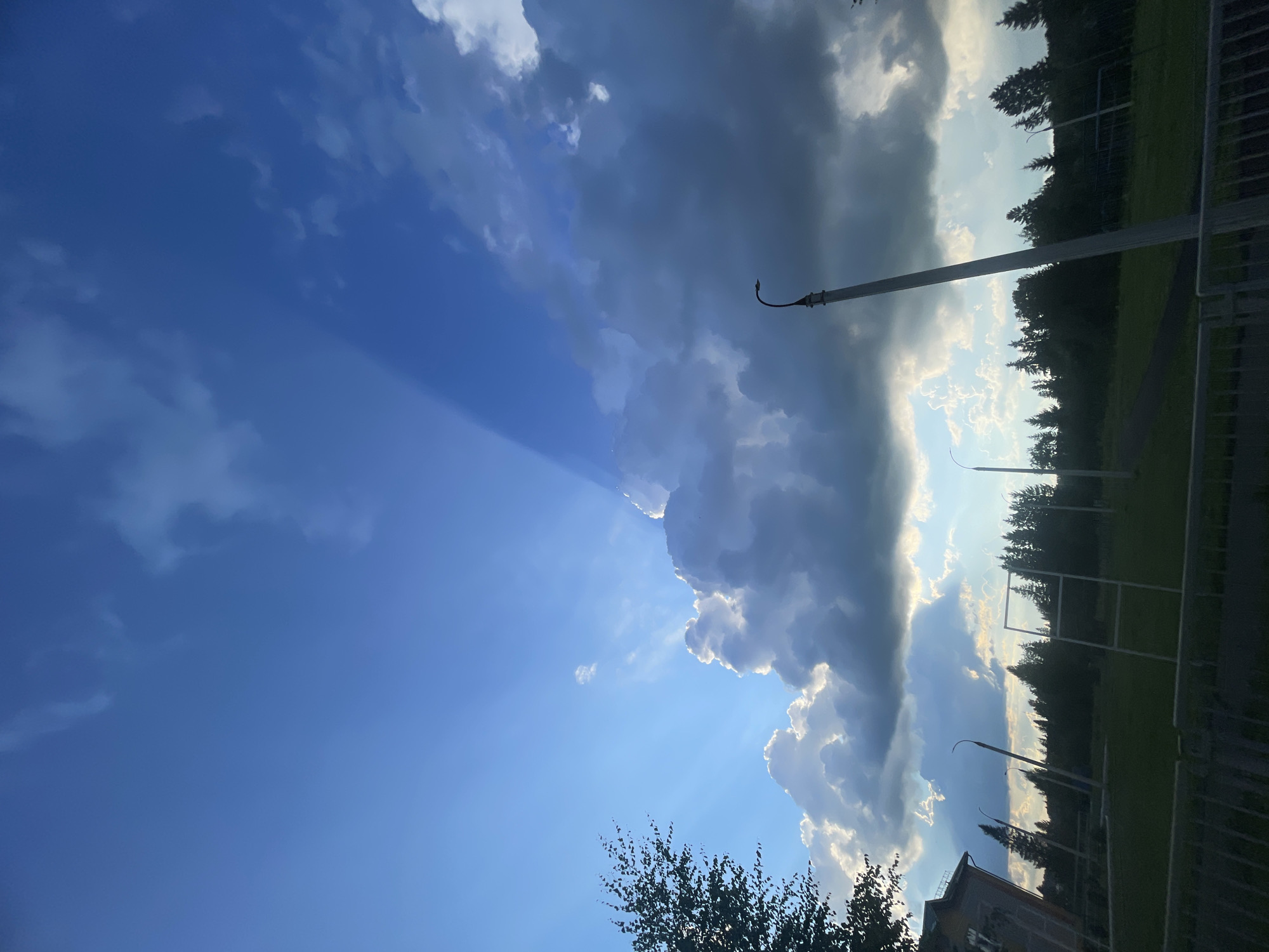 The image captures a dramatic sky over a landscape that appears to be on the outskirts of Podshivalovo, Russia. The sky is dominated by a dynamic interplay of sunlight and clouds. Beams of light, known as crepuscular rays, emanate from behind a large, puffy cloud formation, casting a stark contrast against the deep blue sky. This cloud formation is illuminated from behind, creating a halo effect.
In the foreground, a dark, silhouetted treeline marks the edge of a sparsely vegetated area. Beyond the treeline, the scene transitions to a grassy field dotted with several tall, slender lampposts. One lamppost, positioned slightly off-center to the right, is particularly prominent, leaning at an angle. In the far left of the image, a portion of a building with a textured facade is visible. The overall impression is of an open, somewhat rural or semi-urban environment experiencing a striking natural light phenomenon.
