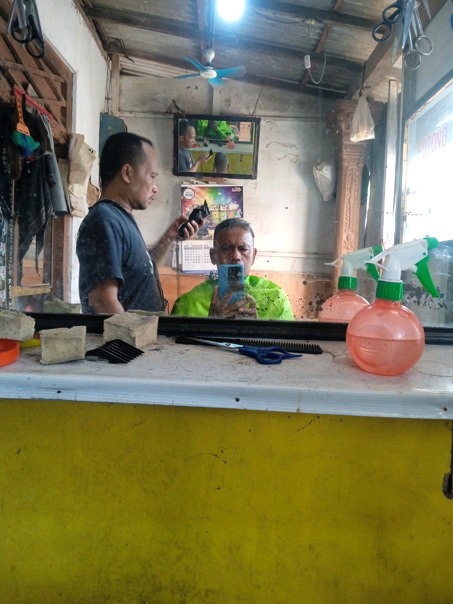 This image depicts the interior of a barbershop in Mangkujayan, Indonesia, as evidenced by the Indonesian word "POTONG" (meaning "cut") visible on a sign. A man, presumably the barber, stands to the left, holding an electric hair clipper and looking into a mirror. In the mirror's reflection, another man, the customer, is seated and holding a smartphone. The customer is wearing a bright green shirt. The barbershop appears to be a modest establishment, with a wooden ceiling, a fan, and various items visible on a shelf and hanging on the wall, including scissors and a brush. In the foreground, on the counter in front of the mirror, are two blocks of what appear to be sponges, a comb, scissors, and two spray bottles, one orange and one pink, both with green nozzles and white triggers. The overall environment is cluttered and shows signs of daily use.
