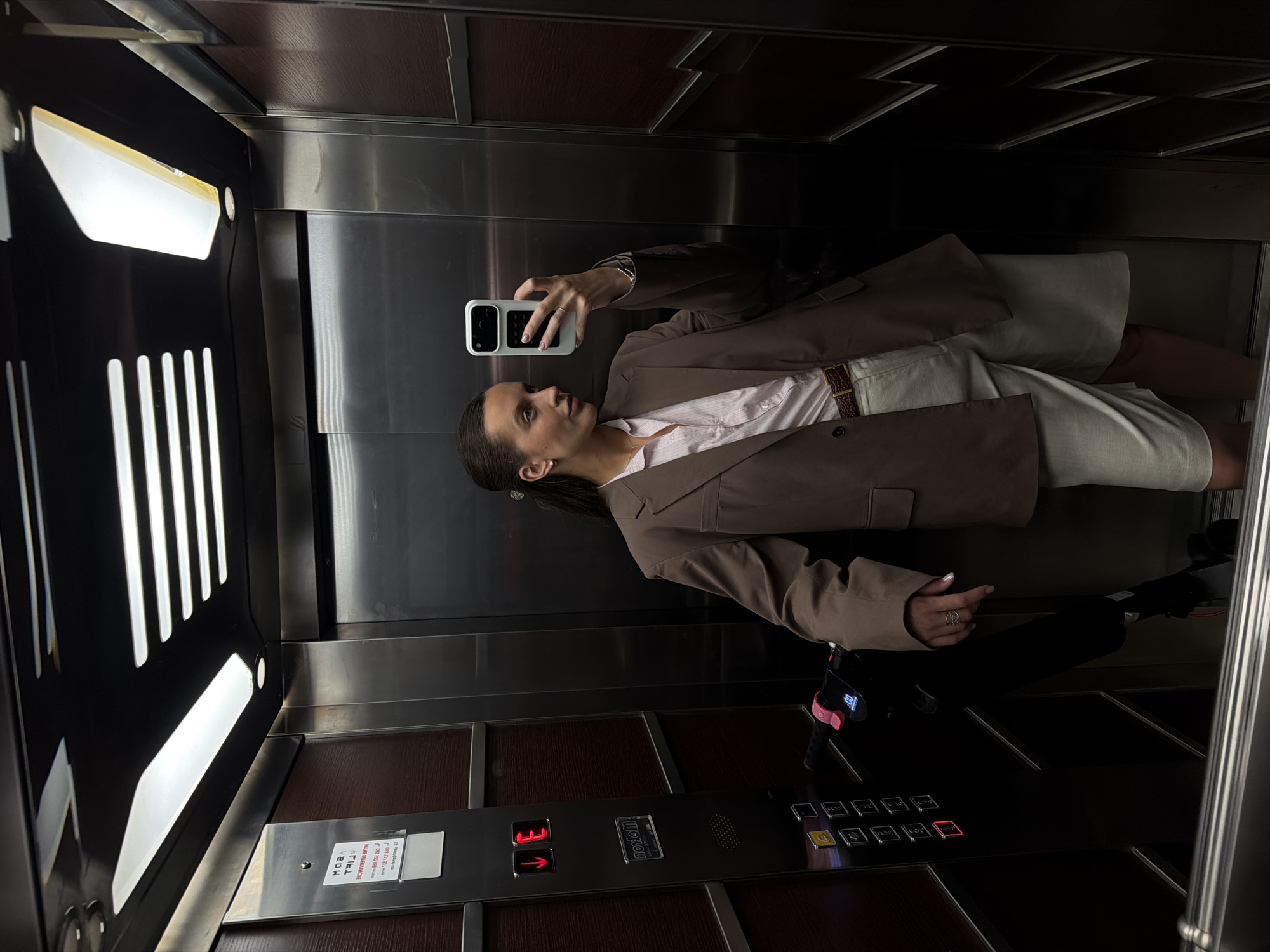 In an elevator, a woman is taking a selfie in the mirror. She is wearing a brown oversized blazer over a light pink collared shirt, and light-colored Bermuda shorts. A brown belt with a textured pattern cinches her waist. She has her hair pulled back in a ponytail and is wearing a ring on her right hand. In her right hand, she holds a white phone, capturing her reflection. To her left, a black electric scooter with pink accents is parked, with its handle bar visible. The elevator walls are a mix of dark wood paneling and brushed stainless steel. Above, fluorescent lights illuminate the space. The elevator control panel is visible on the left, with illuminated buttons. The scene appears to be in an elevator in the city of Budva, Montenegro.