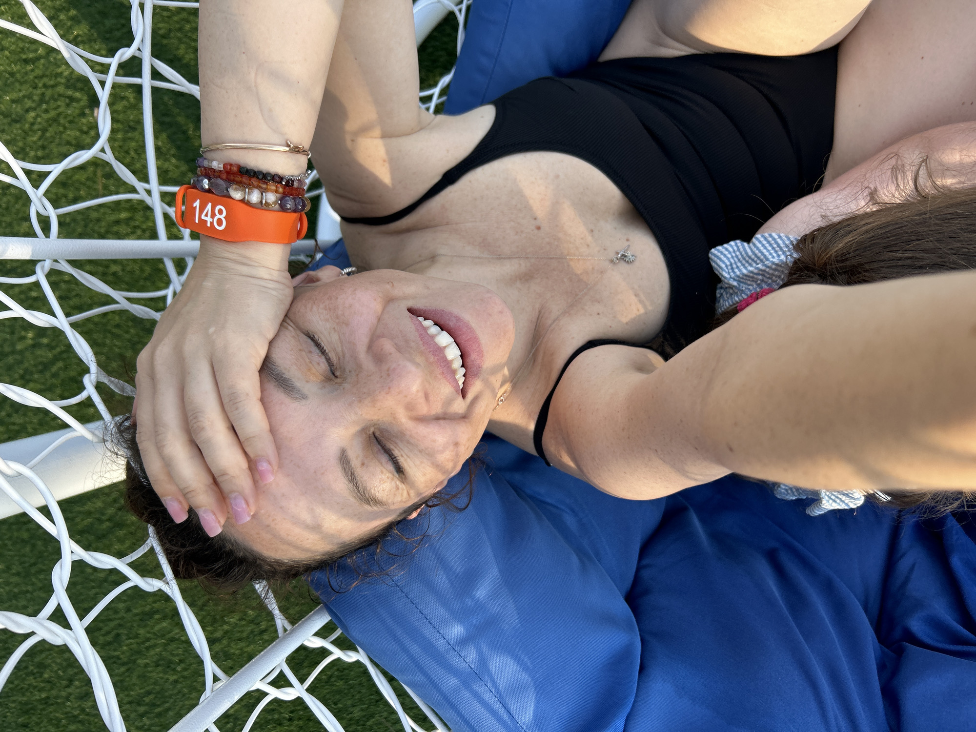 A woman is lying down on a blue cushion, which is part of a white net chair, outdoors. She has her eyes closed and is smiling, with her hand on her forehead. She is wearing a black swimsuit, and several bracelets on her wrist. One of the bracelets is orange and has "148" written on it in white text. The background appears to be a grassy area, and the lighting suggests it is daytime, possibly late afternoon, with warm sunlight. There is a small silver necklace with a charm on her chest. The scene conveys a relaxed and happy emotion.