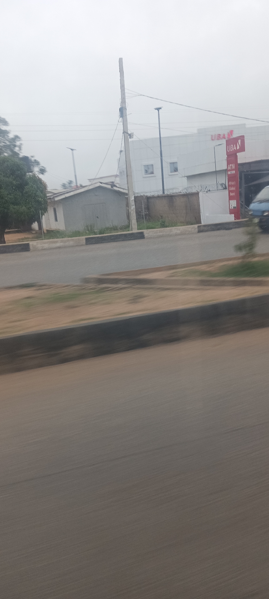 The image presents an outdoor street scene in Jos, Nigeria, captured from a moving vehicle, indicated by the horizontal motion blur, particularly in the foreground and midground. The sky is overcast and hazy, casting a soft, diffused light across the landscape.
In the immediate foreground, a blurred expanse of gray asphalt or ground is visible, followed by a darker strip and another lighter, blurred section, suggesting the vantage point is from a vehicle window looking out. The midground features a paved road with a distinct curb, painted with black and white stripes, running parallel to the direction of motion.
Beyond the road, the background consists of commercial and utility structures alongside greenery. To the left, dense green foliage from a tree is visible, next to a small, light-colored building with a pitched roof, possibly a shed or a small house. A fence appears to separate this property from the roadside.
On the right side, a prominent, modern white building with small, square windows stands out. This building is clearly identified as a bank by a large red sign displaying "UBA." Below it, a smaller red sign advertises "ATM" and the slogan "Africa's Global Bank." A utility pole with various wires stands in front of the bank. A concrete wall topped with barbed wire runs along the bank's perimeter, adjacent to the smaller building on the left. A dark-colored vehicle, possibly a van or small truck, is partially visible and blurred on the far right. Several street lights are also scattered throughout the scene. The image portrays an active urban or suburban environment on a cloudy day.