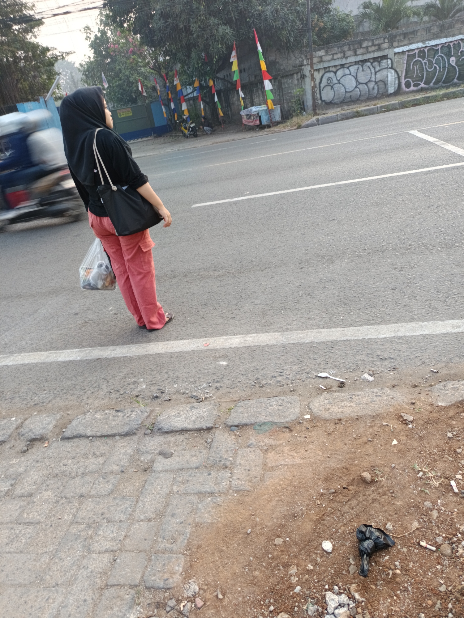 A person wearing a black hijab and black top is standing on the side of a road in Talaga Sari, Indonesia. They are wearing pink pants and dark sandals. The person is holding a black bag in their left hand and a clear plastic bag filled with items in their right hand. A motorcycle is blurred in motion to the left of the person. The road has a white line marking and a curb. To the right of the road, there is a wall with graffiti. The ground beside the road is dirt and gravel, with some scattered trash, including a black plastic bag and cigarette butts. Trees and flags are visible in the background.
