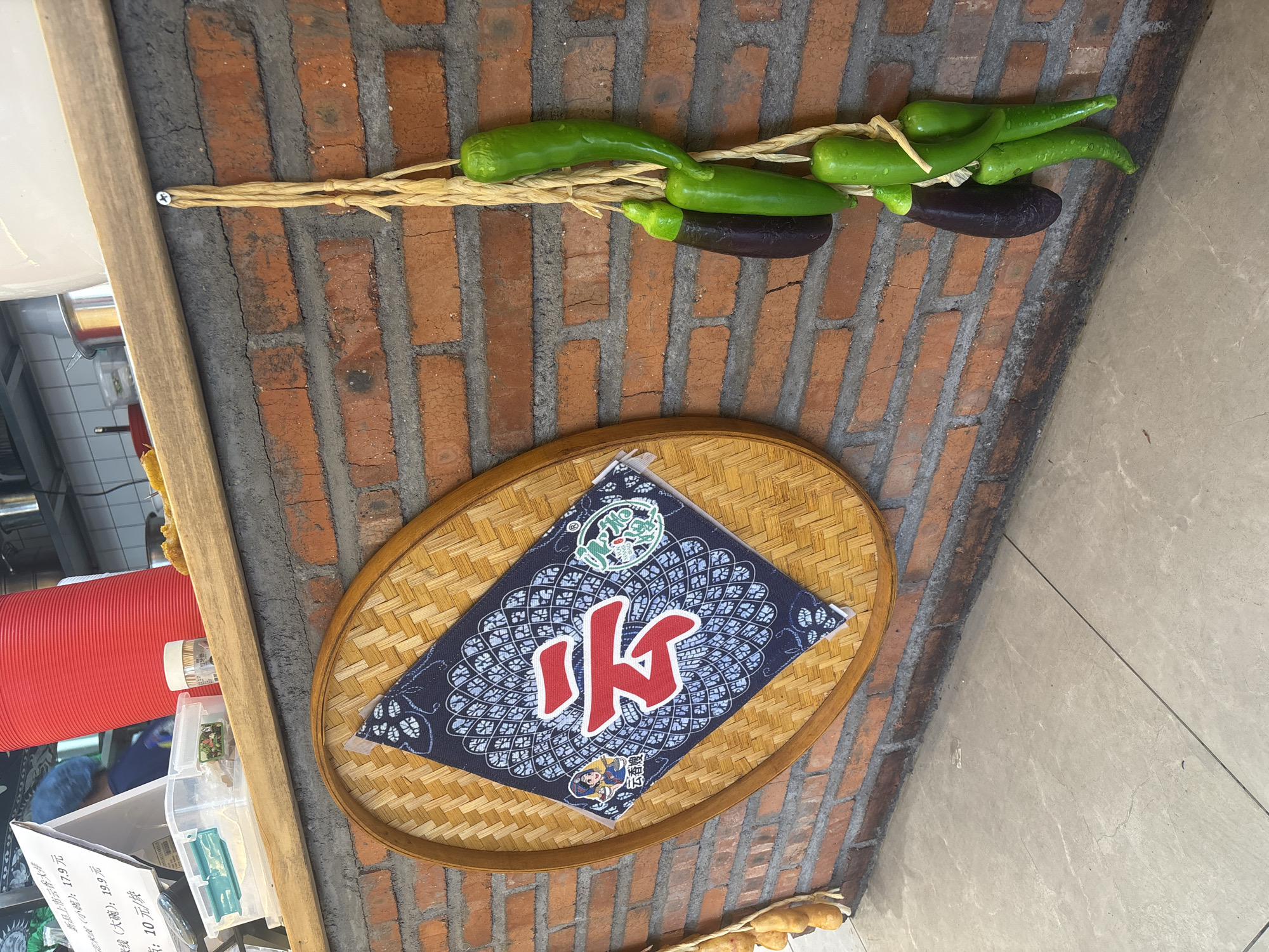 The image captures a close-up of a brick wall in a food establishment, likely a restaurant or food stall, in Suzhou Industrial Park, China. The wall is made of weathered red bricks with grey mortar. Hanging on the wall, from a rough rope tied to a screw, are several green bell peppers and two dark purple eggplants, arranged vertically. To the left of the hanging vegetables, a round, woven bamboo mat is affixed to the wall. Centered on the mat is a dark blue fabric with a white paisley-like pattern, upon which a large red Chinese character "云" (yún, meaning cloud) is prominently displayed in the center. To the left of the character, a small circular emblem features a cartoonish illustration of a woman in traditional Chinese attire and a smaller, circular green emblem with Chinese text. The overall impression is one of rustic decoration within a food service setting. In the background, out of focus, are glimpses of a wooden counter, a bright red cylindrical object, and some plastic containers.