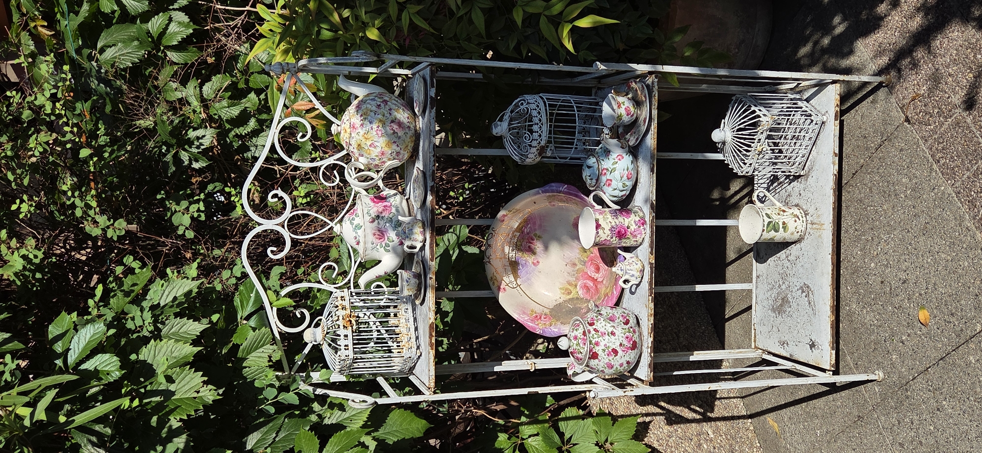 The image shows a white, multi-tiered metal shelf unit set up outdoors against a backdrop of lush green foliage. The shelves are filled with various decorative items, primarily porcelain teapots, teacups, and saucers, all adorned with floral patterns. There are also several white decorative birdcages and a large decorative platter with a floral design. The shelf unit and its contents are in a state of disrepair, with visible rust and peeling paint. The ground in front of the shelf is paved with grey and speckled tiles, with some fallen leaves scattered around. The lighting suggests a sunny day, with distinct shadows cast by the shelf unit and its objects. The environment appears to be a garden or patio area. There are no people or animals visible in the image. The specific location in Strasbourg, France cannot be determined from the visual information provided.