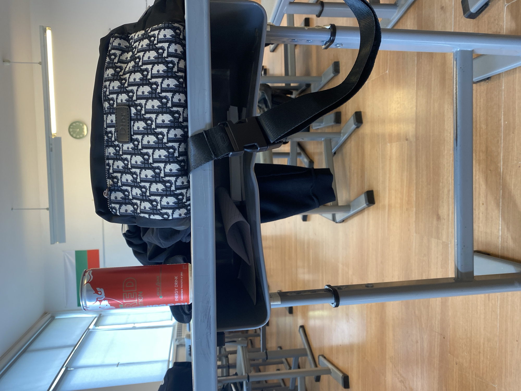 The image shows a classroom setting, likely in Varna, Bulgaria, suggested by the presence of the Bulgarian flag in the background. The primary focus is on a grey school desk with an attached seat. A Dior-branded backpack is hanging from the back of the chair. A partially visible red can of Red Bull energy drink sits in the desk's storage area below the seat. The classroom appears to be well-lit, with natural light coming through the window and supplemented by fluorescent lighting. The floor is wooden, and other desks are visible in the background, creating a sense of depth. The desks seem to be aligned in rows. No people are directly visible.