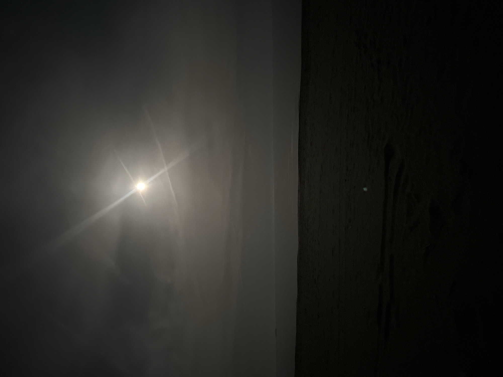 The image depicts an outdoor scene, likely at night, in Donji Štoj, Montenegro. A light source, possibly the moon or a streetlamp, is visible on the left, obscured by mist or fog. The light creates a bright glow with rays emanating outwards. The left side of the image appears to be a wall or flat surface catching the light. To the right of the wall, the scene transitions into deep shadow, revealing a rough or textured surface which may be another wall or vegetation.