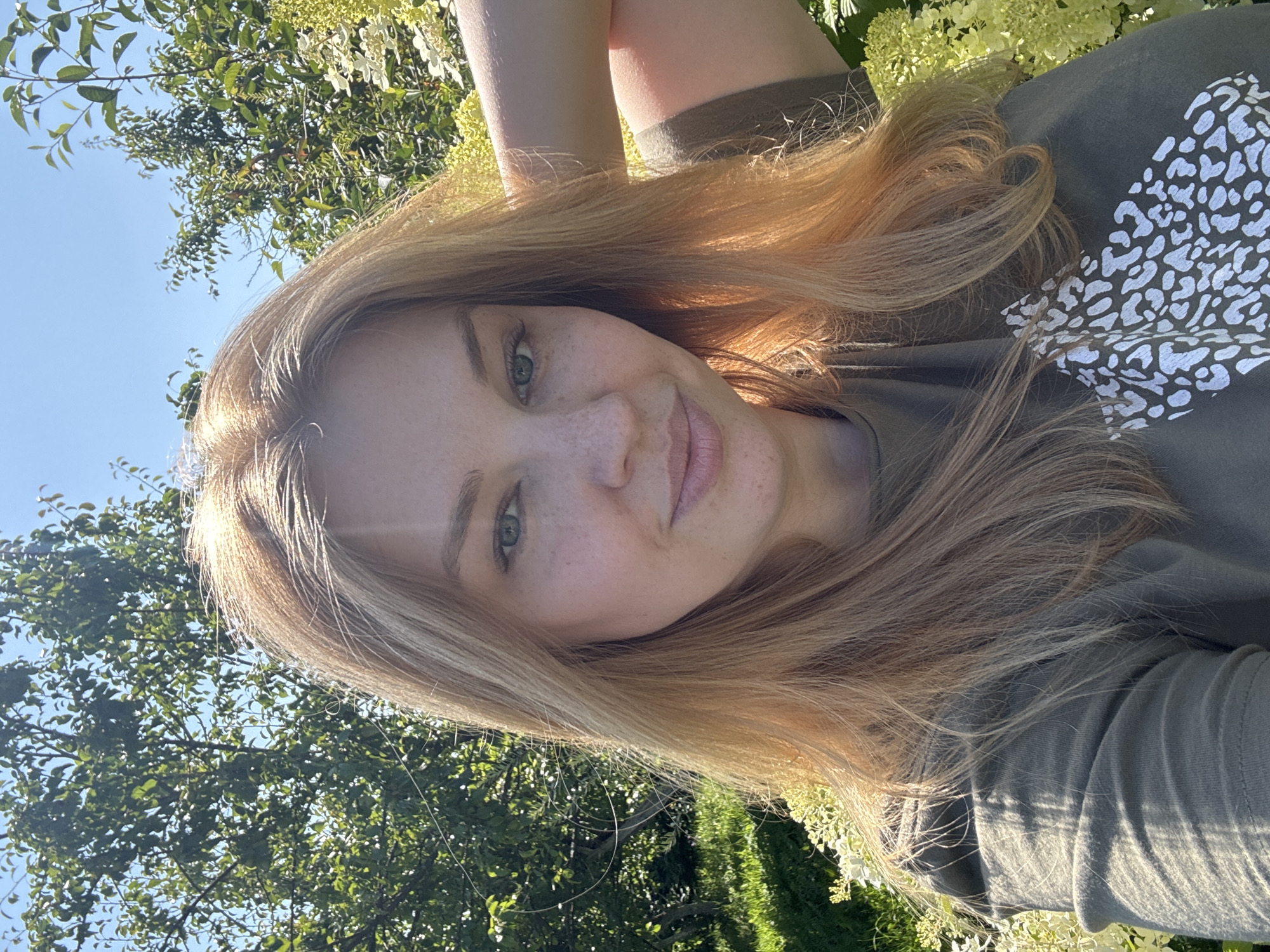 A Caucasian woman with shoulder-length blonde hair and fair skin with freckles is taking a selfie outdoors. She is wearing a green t-shirt with a white leopard print design. She has her right arm raised and bent behind her head, with her hand resting on her hair. The background shows lush green trees, foliage, and a bright blue sky. The image appears to be taken in a garden or park setting. The lighting suggests it is daytime, possibly late afternoon, with sunlight filtering through the leaves. The overall scene conveys a casual and relaxed atmosphere.