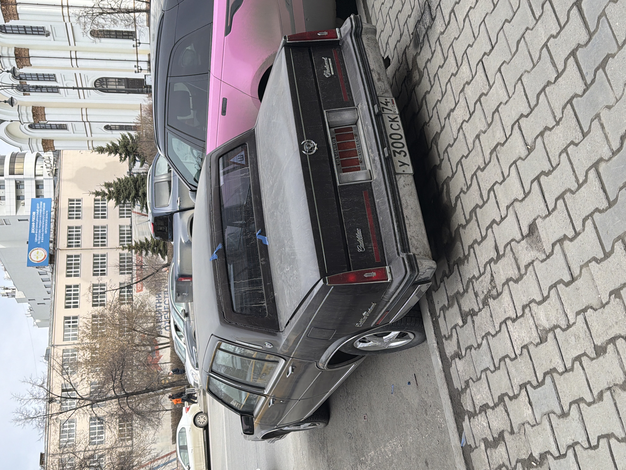 The image shows a black Cadillac parked on a street, possibly in Yekaterinburg, Russia, as suggested by the Russian license plate "Y 300 CK 74." The car is parked on a sidewalk made of gray paving stones. Adjacent to the Cadillac is another vehicle, a pink SUV. In the background, there are several buildings, including one with a blue sign indicating "ДОСААФ РОССИИ," a Russian organization. Bare trees are visible along the street. The image appears to be taken during the day.
