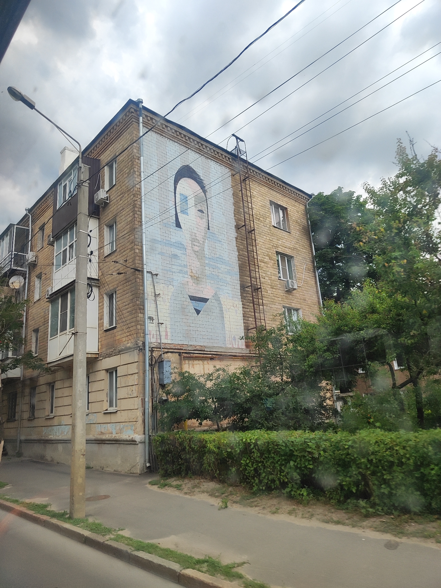 A mural of a person's face is painted on the side of a brick building. The building is multi-story with windows and balconies. There is a street light in the foreground and trees and bushes surround the building. The sky is cloudy.