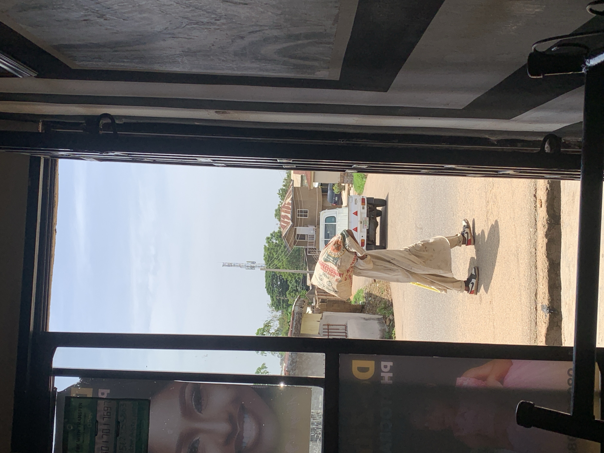 The photograph shows an outdoor street scene, viewed through the dark frame of an interior doorway or large window. The day is bright and sunny with a clear sky, indicative of midday or early afternoon.
On a paved street, a person walks from right to left, expertly balancing a large white sack on their head. The sack completely covers their head and upper torso. They are dressed in a light-colored, loose-fitting garment and wear black, white, and red sneakers. Text visible on the sack includes: "GOODS," "CONFECTIONERY GREAT FLOUR," "50 KG NET," and "MADE IN NIGERIA PLC."
Further down the street to the right, a white pickup truck is visible. The background features several modest buildings, including a house with a brown roof, interspersed with green trees and vegetation. A tall communication tower stands in the distant background.
In the immediate foreground, the interior ceiling or wall features a black and light grey geometric pattern. A glass pane on the lower left reflects part of an advertisement, showing a smiling face and partial text such as "PAYMENTS," "TRANSFER," "OPay," and the numbers "17697."