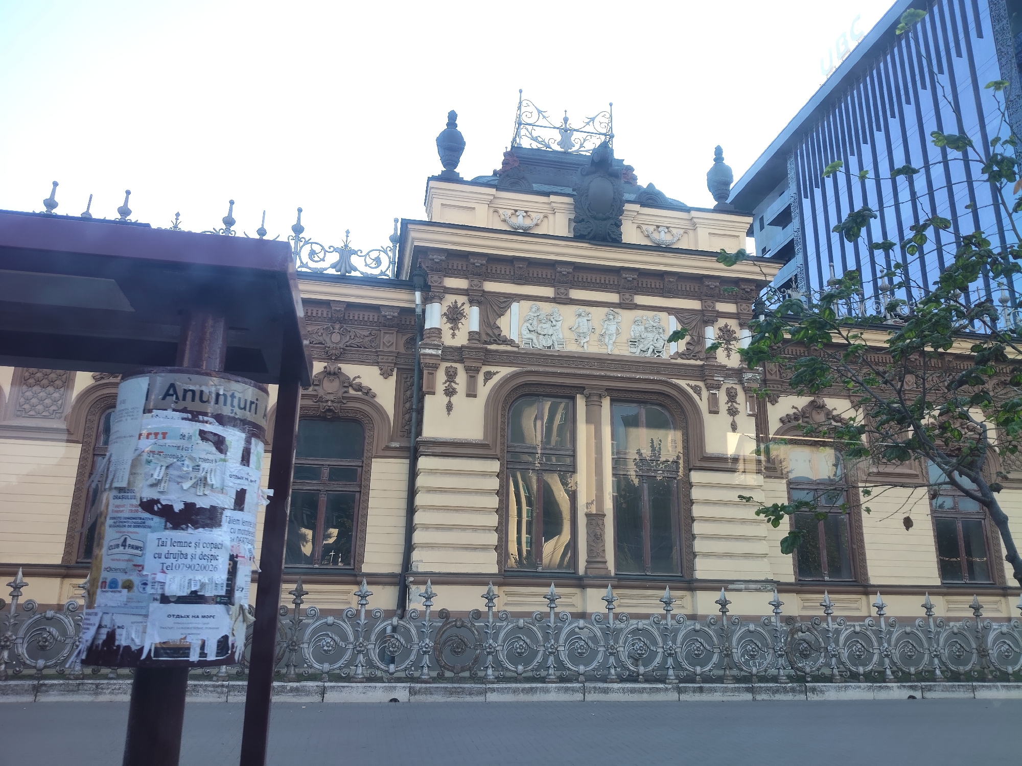 The image captures the facade of a historical building in Chisinau, Moldova, with a modern glass-fronted building visible to the right. The historical building features ornate architectural details, including intricate carvings and decorative elements around the windows and roofline. It is painted in a light yellow hue with darker brown accents. In the foreground, a cylindrical advertising pole is covered in various torn posters, some bearing text in Romanian. A metal fence with decorative circular motifs runs along the front of the historical building. A tree with green leaves partially obscures the right side of the historical building and the modern glass building, which has vertical blue elements. The sky is overcast and bright, suggesting daytime.