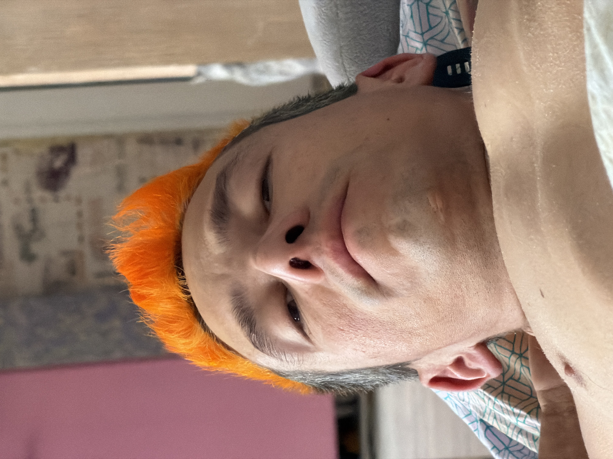 The subject is an adult male positioned reclining on his back in an indoor residential setting in the city of Chaikovsky, Russia. He is facing upwards towards the camera, with his head slightly tilted and his gaze directed upwards. His hair is a prominent feature, dyed a vivid, bright orange and styled upwards in a spiky fashion on the crown, contrasting with shorter, graying hair visible on the sides of his head.
His facial features include thick, dark gray eyebrows, dark eyes partially visible and looking upwards, and a prominent nose with visible nostrils. His lips are closed in a relaxed state. A faint stubble is visible on his chin and jawline. His skin appears fair with visible pores and minor textural irregularities. Both ears are partially visible, with the left ear (viewer's right) showing more detail.
The subject is bare-chested, revealing a portion of his upper chest and shoulders, where a small, raised mole or skin tag is present on the upper left chest area. Faint body hair is visible on his chest. A dark-colored wristband or watch strap is partially visible behind his right ear, suggesting it is worn on his right wrist.
He is resting on a light-colored, geometrically patterned pillowcase or sheet, featuring light blue/teal lines forming a grid-like design, visible beneath his head and to the viewer's left. A soft, gray fabric object, likely another pillow or blanket, is situated to his right.
The background consists of blurred interior elements: a plain, dull pink/magenta wall to the viewer's left, a darker textured wall above it, and a light-colored vertical structural element (such as a door frame) to the viewer's right, with another blurred wall behind it. The lighting is soft and even, suggesting ambient indoor illumination. No direct action or interaction is depicted beyond the subject's gaze towards the camera.