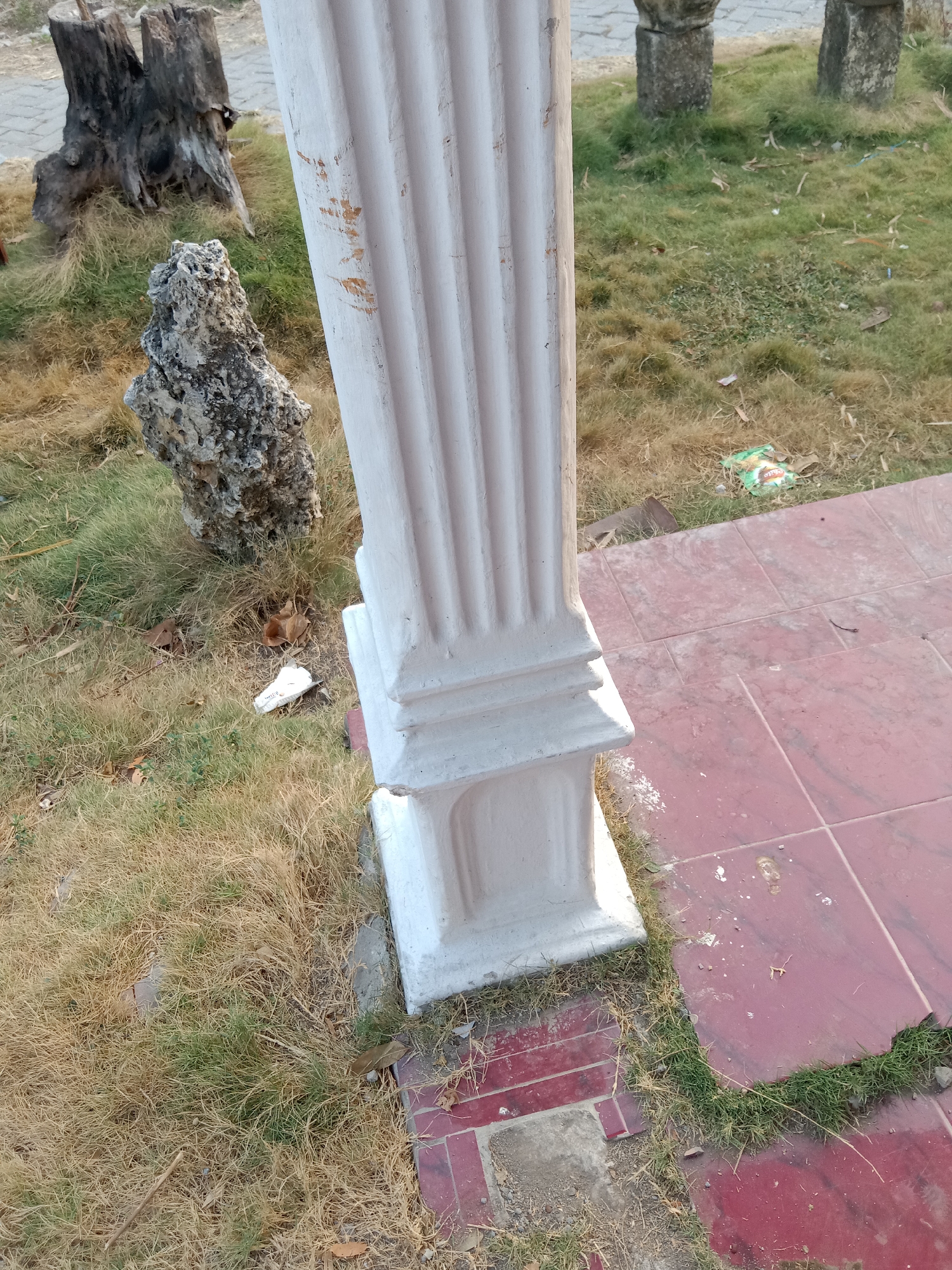 The image depicts an outdoor scene in Pacing, Indonesia, focusing on a white, fluted pillar standing next to a reddish-tiled pathway. The pillar has a square base and a detailed, tiered pedestal. To the left of the pillar, the ground is covered in a mix of dry, yellowed grass and patches of greener grass, with some scattered leaves and debris. A large, grey, rough-textured rock sits in the grass, and behind it are the weathered remains of two tree stumps. In the background, slightly out of focus, are glimpses of more stone structures and more of the grassy area. There are no people visible in the image.