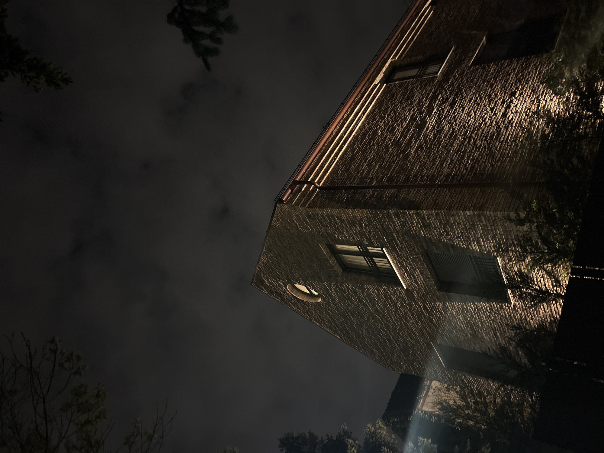 A low-angle night shot depicts a brick building with illuminated walls. Two windows are visible, along with a circular vent. The top edge of the building shows several lines of trim. The sky is cloudy and dark. Trees are visible in the foreground. It is not possible to determine the location is Sofia, Bulgaria based solely on the visual information.