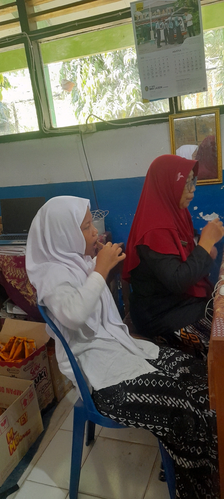 The image depicts an indoor scene, likely a classroom or office, with two individuals present. The person on the left is a young female wearing a white hijab and a white long-sleeved shirt. She is seated in a blue chair and appears to be holding something to her mouth with her right hand, possibly eating. To her right, another individual is wearing a red hijab and a black top with glasses. This person is also seated and seems to be interacting with something in their hands.
In the background, a window with green trim is visible, revealing trees and foliage outside. A calendar for 2026 hangs on the wall above the window, displaying the months of July and August. A framed picture or mirror is also present on the wall to the right. Various items are scattered around the room, including boxes, a laptop, and what appears to be food packaging on the floor. The flooring is tiled. The overall environment suggests a casual or educational setting within the city of Tahunan, Indonesia, given the calendar's mention of "BKK LASAM," a known entity in the region.