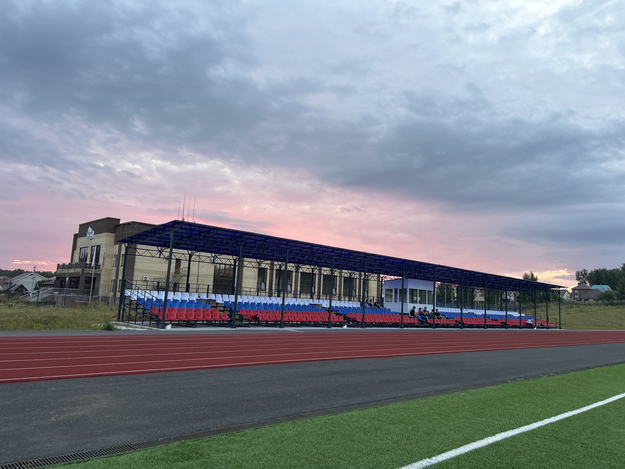 The image captures an outdoor sports stadium in Noviy, Russia. The foreground features a red running track and a green artificial turf field. Behind the field are tiered bleachers with seats colored red, white, and blue, arranged in a repeating pattern. Several people are seated on the bleachers, some conversing. To the left of the bleachers is a brick building with a blue roof structure and several windows. The sky above is filled with dramatic clouds, showing a gradient from dark grey to a soft pink and orange hue, indicative of a sunset. The overall scene depicts a sports facility under a colorful evening sky in a semi-rural setting, with a few houses visible on a distant hill.