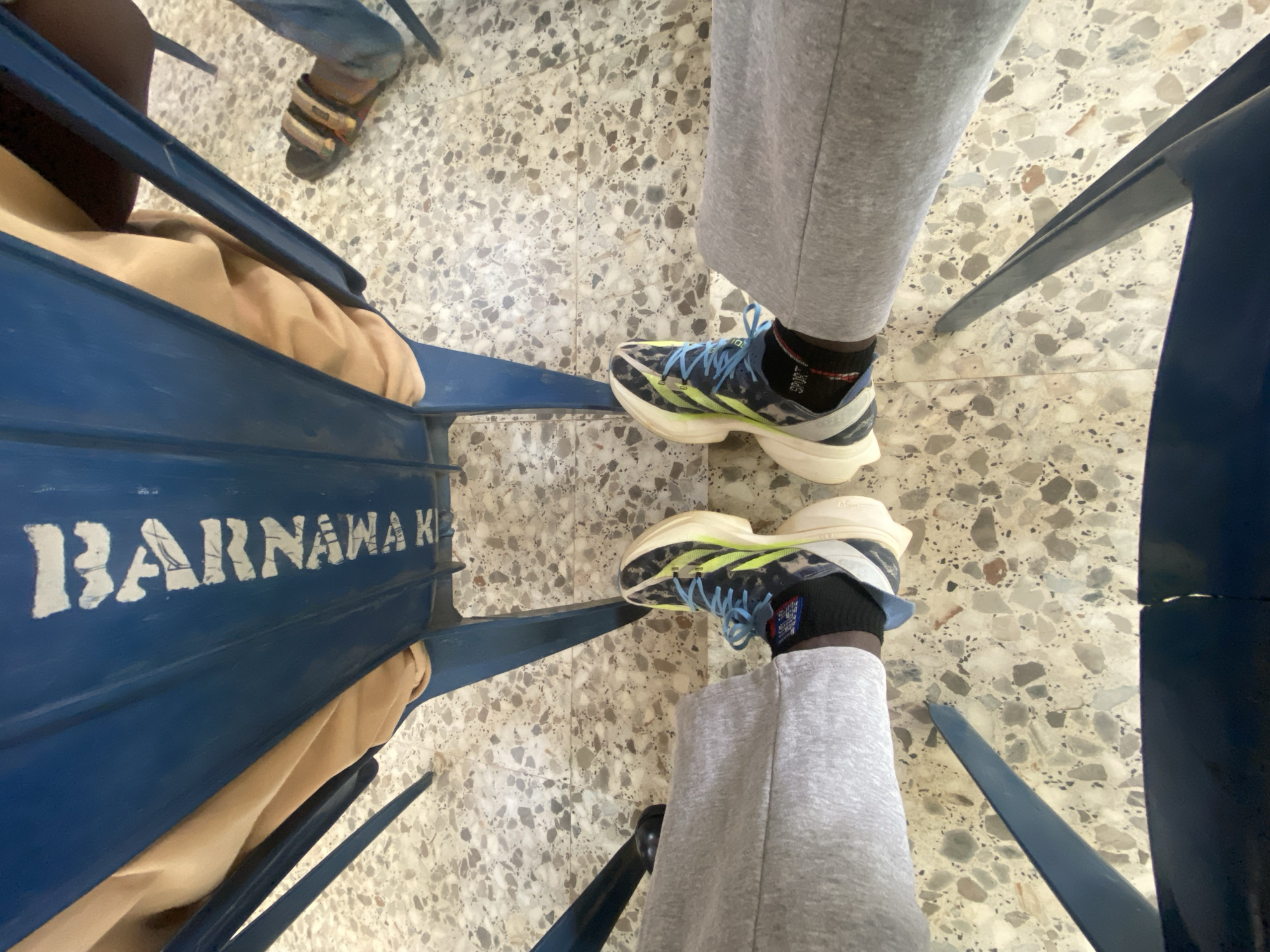 The image shows a view from above, looking down on three people seated in blue metal chairs on a terrazzo floor. One person is wearing sandals, another is wearing grey sweatpants and running shoes with blue laces and a white sole. A tan-colored cloth is draped over one of the blue metal chairs, labeled "BARNAWA K..." in white letters. Based on the label, the location is inferred to be Barnawa, Kaduna, Nigeria.