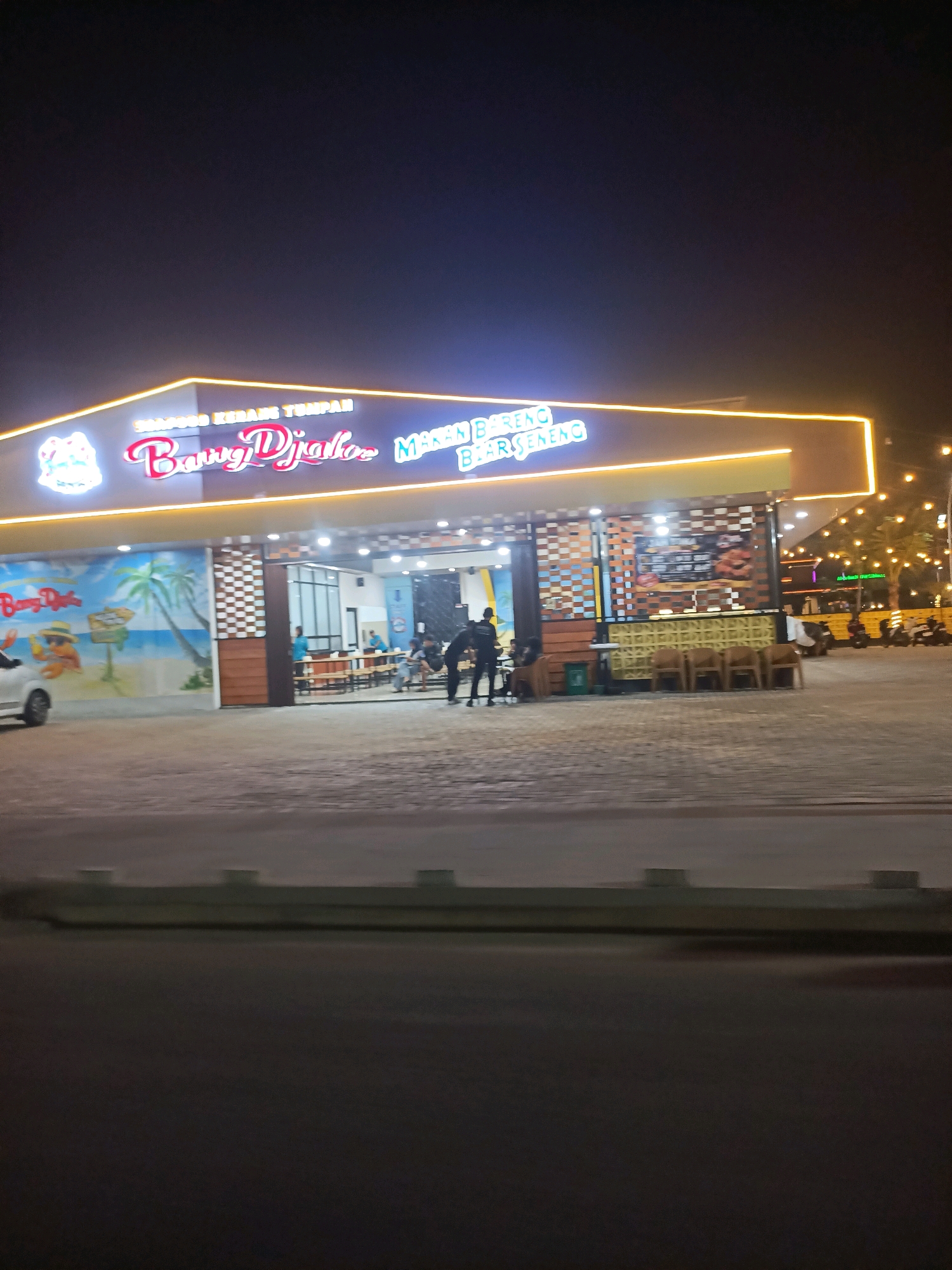 The image captures a nighttime scene outside a building identified by illuminated signage that reads "SEAFOOD KERANG TUMPAH". Below this, another sign, "BannyD'jako", is prominently displayed in cursive font. To the right of this, text in capitalized letters appears to read "MAKAN BARENG BIAR SENENG". The building features a wide facade with several open doorways leading into an interior area where tables and chairs are visible, along with people seated. The exterior wall on the left is decorated with a mural depicting a beach scene with palm trees and a figure. A white car is parked to the left of the mural. The overall environment appears to be an outdoor dining or entertainment venue. A street or roadway is in the foreground, and beyond the building, more lights are visible, suggesting an urban or commercial setting. The location is identified as Telagamurni, Indonesia.