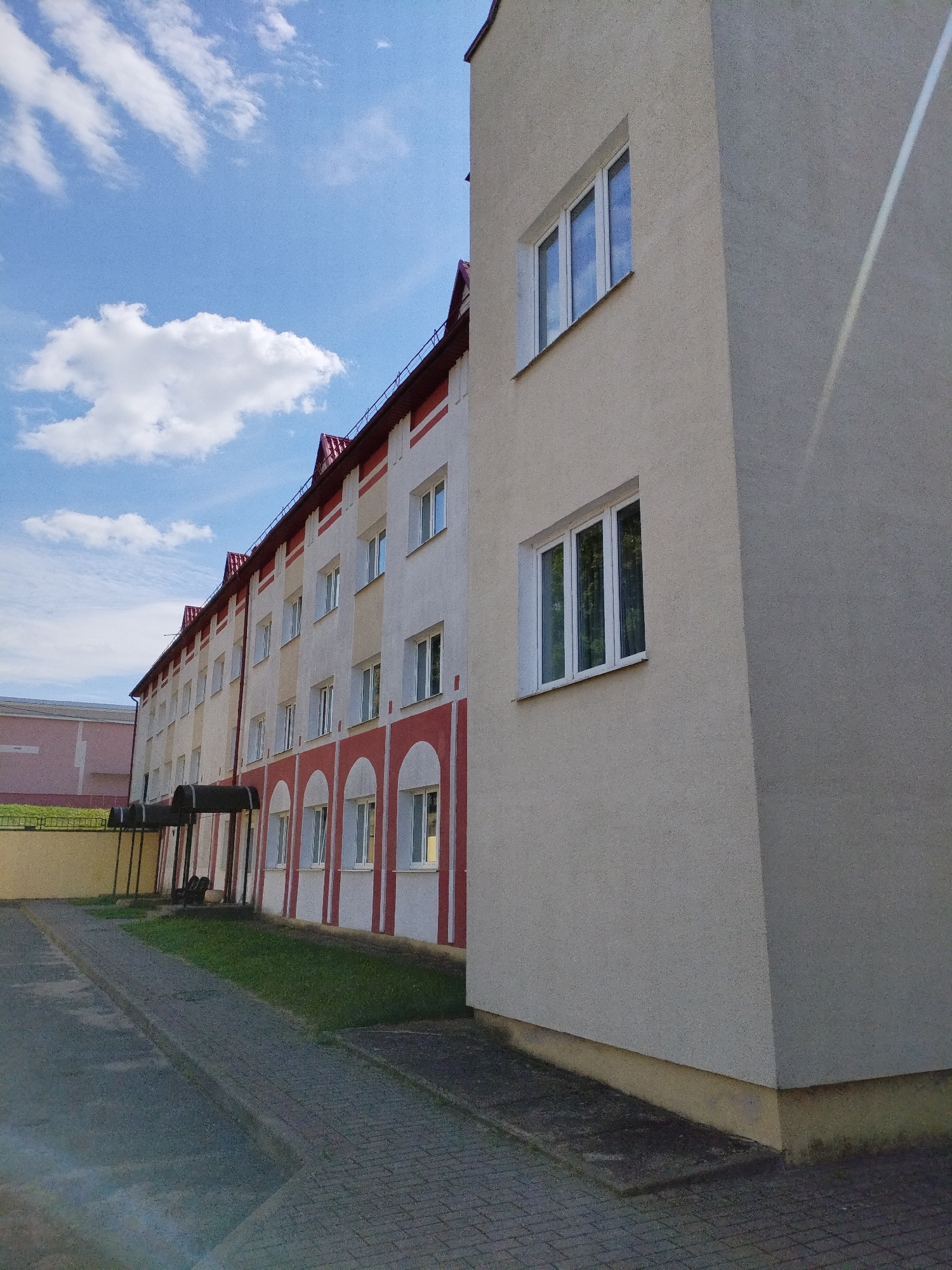 The image shows a multi-story building with a beige facade and red and white trim. The building has several windows with white frames. A paved walkway runs alongside the building, bordered by a strip of grass. In the background, a pink building is visible. The sky is blue with scattered white clouds. The scene is located in Bialynichy, Belarus.