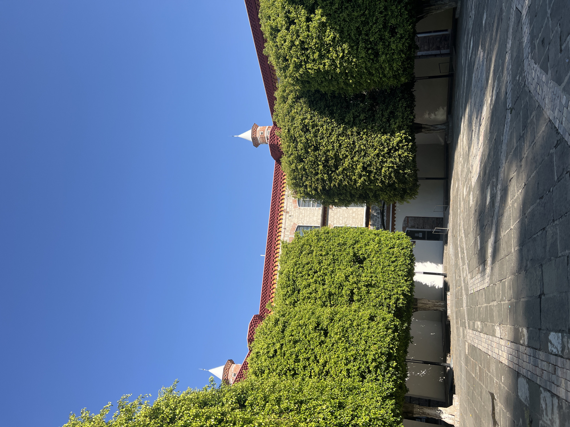The image captures a view of a building in Puebla, Mexico, characterized by a historical architectural style. The building features stone construction, a red-tiled roof, and decorative elements. Green, manicured shrubbery lines the facade, giving the impression of a garden integrated into the building's structure. A courtyard with stone pavement is visible, suggesting a public or semi-public space. The clear, blue sky indicates a sunny day. No people are visible in the frame.