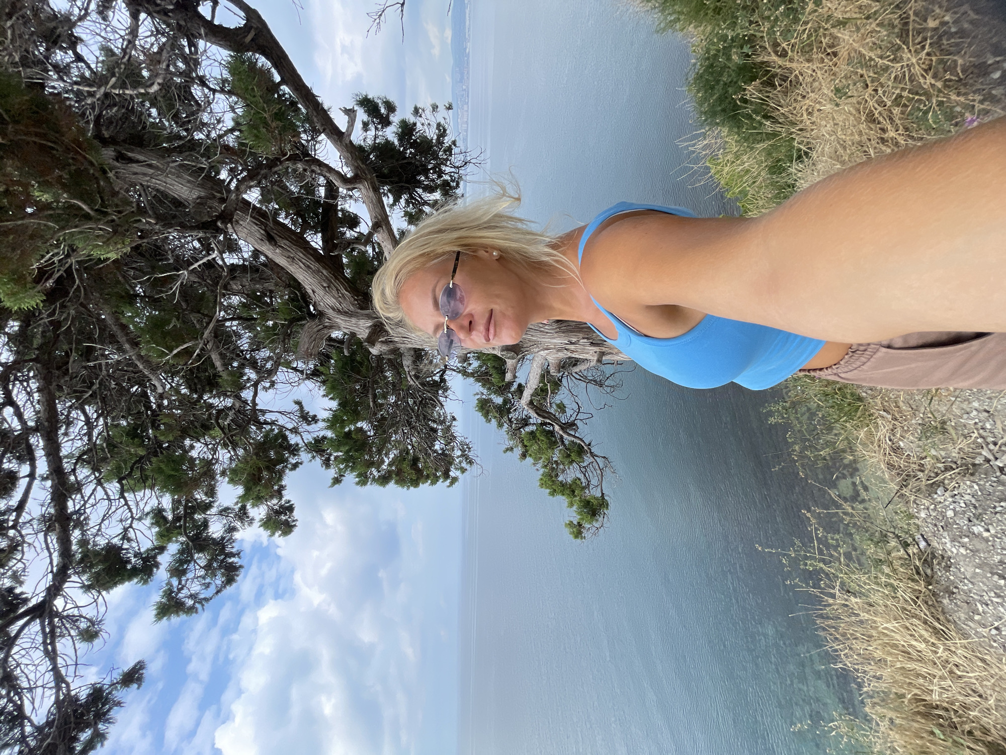 A blonde woman wearing a blue tank top and brown pants takes a selfie on a cliff overlooking the sea. She is positioned on the right side of the frame, with her arm extended towards the camera. A large, gnarled tree with green foliage dominates the upper portion of the image, its branches reaching out over the water. The sea is calm and a deep blue, with a faint line of distant mountains or buildings visible on the horizon. The sky is partly cloudy. The foreground shows dry grass and gravel. The scene is likely located on the coast of Novorossiysk, Russia, possibly near Mys Utrish, a nature reserve known for its unique juniper forests and coastal cliffs.