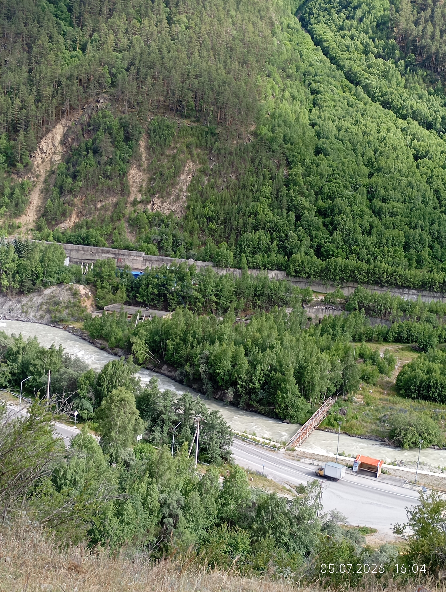 The image depicts a scenic mountain landscape near the city of Neutrino, Russia. A wide, muddy river flows through a valley, flanked by steep, tree-covered slopes. A road runs alongside the river, and a small orange bus stop shelter is visible. A truck is parked near the bus stop. A metal pedestrian bridge spans the river in the distance. The overall impression is one of a remote and rugged natural environment, with signs of human infrastructure present.
