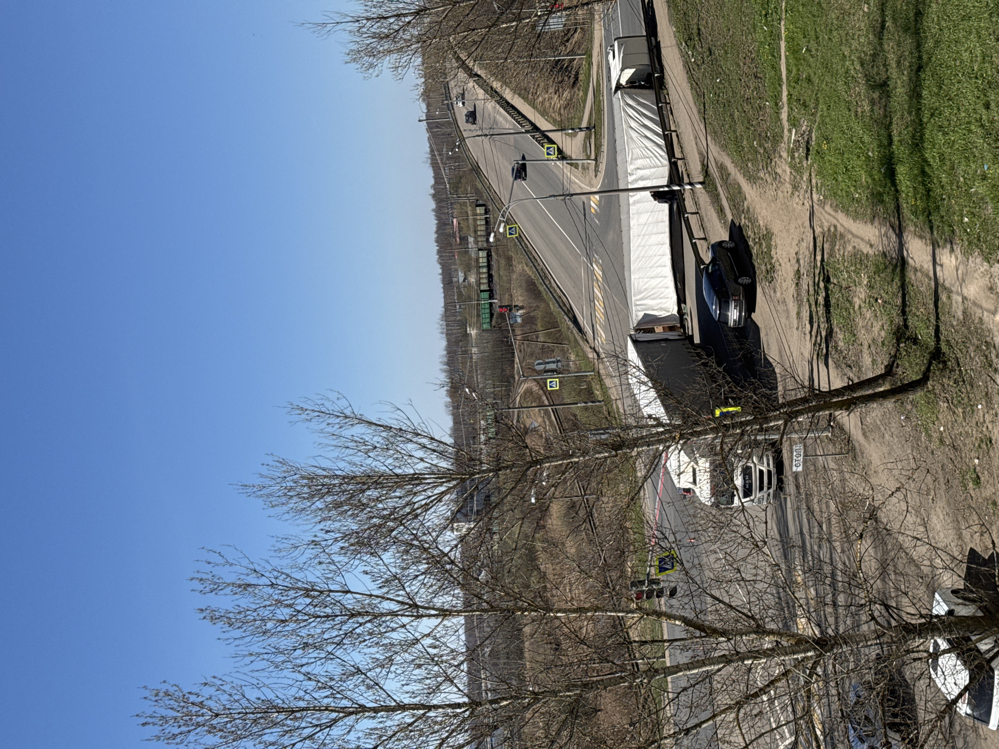 The image depicts a road intersection in Ново-Никольское, Russia. A large white truck is parked on the right side of the road, with a dark car behind it. A second truck is parked further down the road. A railway line runs parallel to the road. A blue sky is visible above, partially obscured by bare tree branches in the foreground. Traffic lights and pedestrian crossing signs are present. The area beside the road is a mix of dirt and grass.