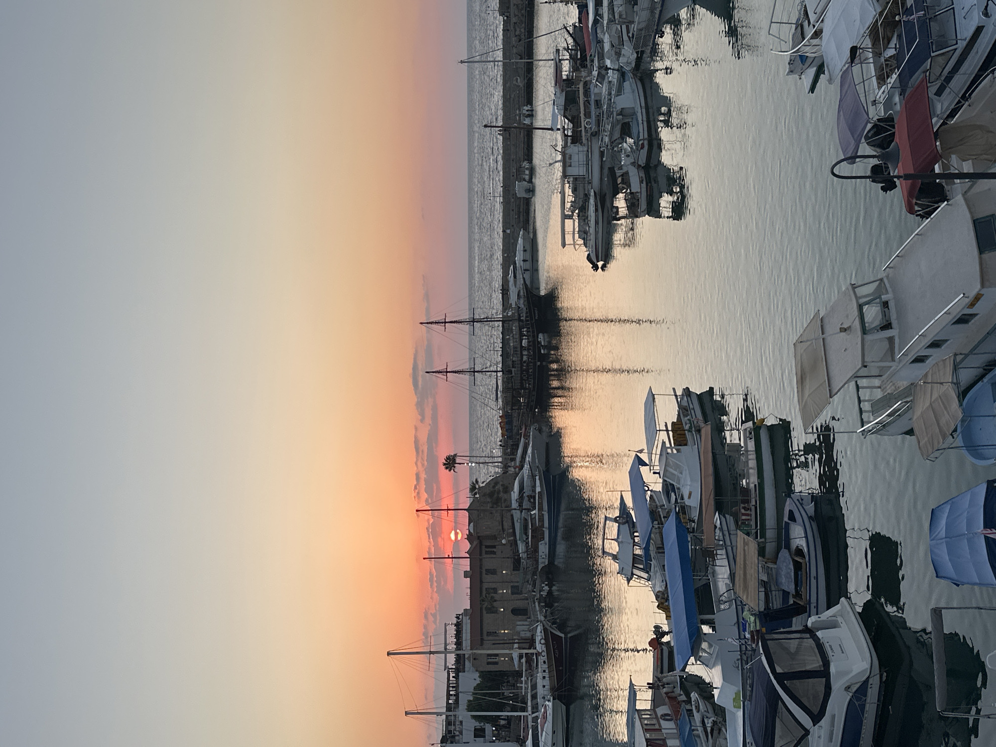This is a medium-shot, vertical photograph of a harbor at sunset in Kyrenia, Northern Cyprus. The sun is a small, orange disc low on the horizon, casting a warm glow over the scene. The sky transitions from a pale blue at the top to a soft orange and pink gradient near the horizon.
The harbor is filled with numerous boats of various sizes, predominantly docked. In the foreground, several smaller motorboats with protective coverings are visible. Further into the harbor, a mix of motorboats and sailboats are moored. Notable are several larger sailboats with tall masts, some appearing to be older or more traditional in design, including a prominent dark-hulled vessel on the right.
Behind the boats, a waterfront promenade is lined with buildings, some of which appear to be historical structures made of stone. A palm tree is visible near the buildings. The water in the harbor is calm, reflecting the colors of the sunset and the shapes of the boats. In the distance, a breakwater or pier extends into the sea, and the gentle waves of the Mediterranean Sea can be seen. There are no visible people in the image. The overall atmosphere is tranquil and picturesque.