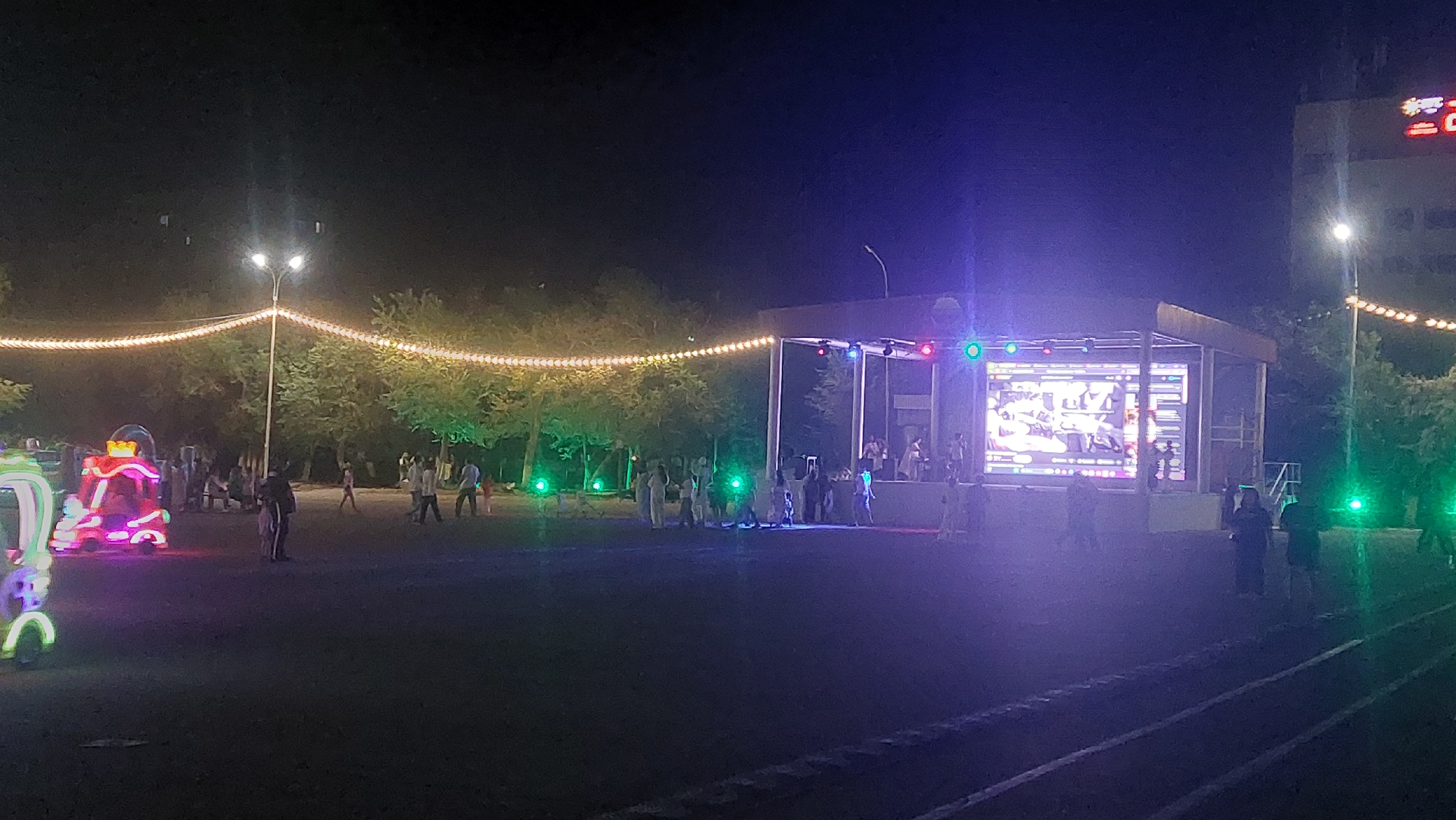 This nighttime outdoor image depicts a scene likely in Priozersk, Kazakhstan, featuring a temporary stage with a large LED screen broadcasting an event. The stage is illuminated with colorful lights, and several people are visible on and around it, some appearing to be performers or event staff. In the foreground, a group of people, including adults and children, are gathered, suggesting an audience or attendees. To the left of the stage, a brightly lit amusement ride or attraction, adorned with neon lights, is present, attracting attention. String lights draped between trees create a festive atmosphere. The environment appears to be a park or an open area within the city, with trees forming a backdrop and a building visible in the far right background. The ground is a dark, paved surface, possibly a recreational area or a street.
