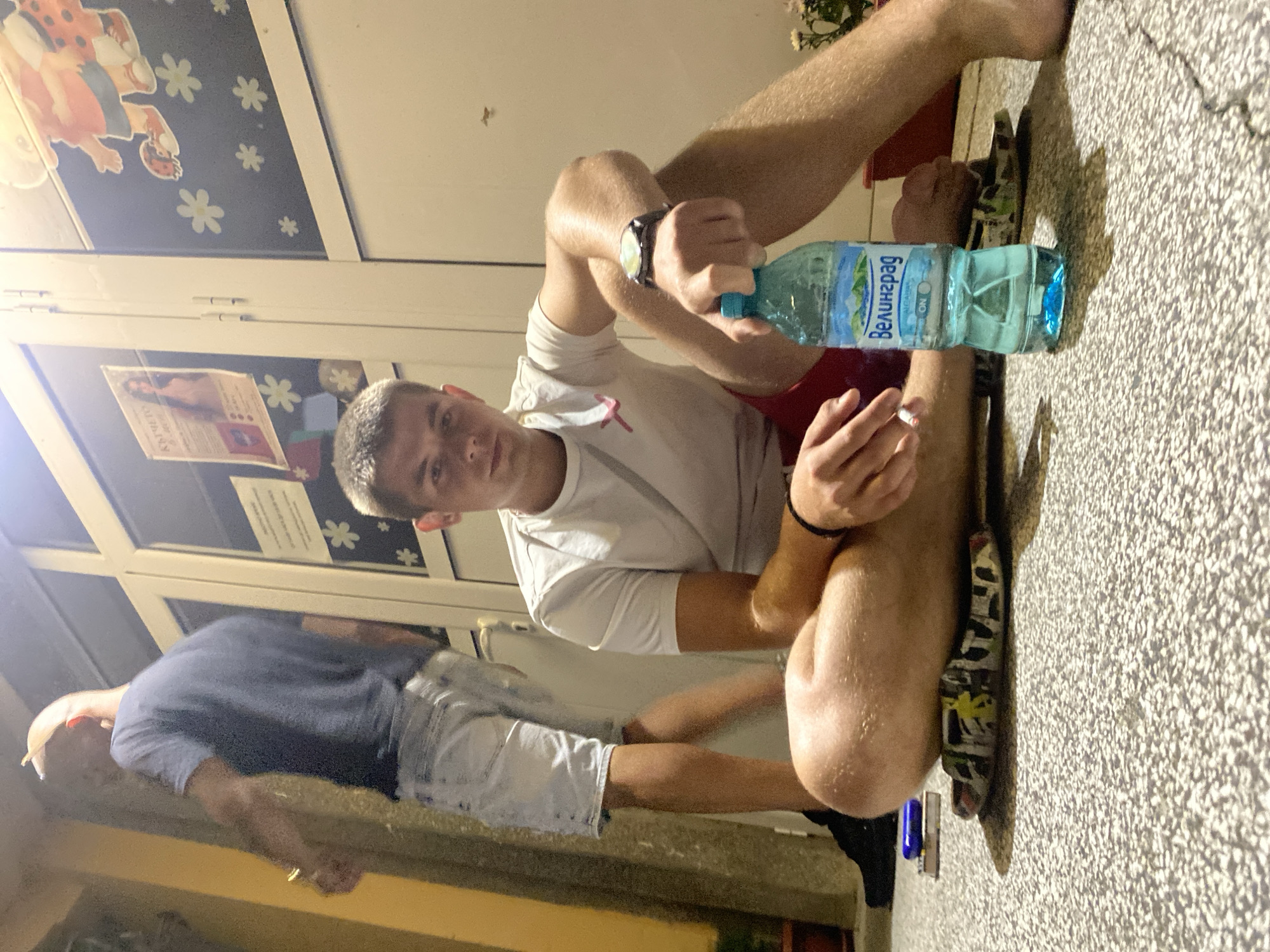 The image depicts two individuals near a building entrance with large glass doors. The person in the foreground is a young man with short brown hair, wearing a white t-shirt and red shorts. He is sitting on the ground, with his legs crossed and resting on a pair of patterned flip-flops. In his right hand, he holds a bottle of water, and in his left hand, he is holding a lit cigarette. A watch is visible on his left wrist. The background shows another man standing, wearing a dark blue t-shirt and light-colored shorts, with his back to the camera. Posters are attached to the glass doors behind them. The ground appears to be paved with gravel. The scene suggests an informal outdoor gathering in a public space. The presence of the building's entrance and the posters could indicate a location within the city of Hirevo, Bulgaria, though this cannot be definitively confirmed from the visual information alone.