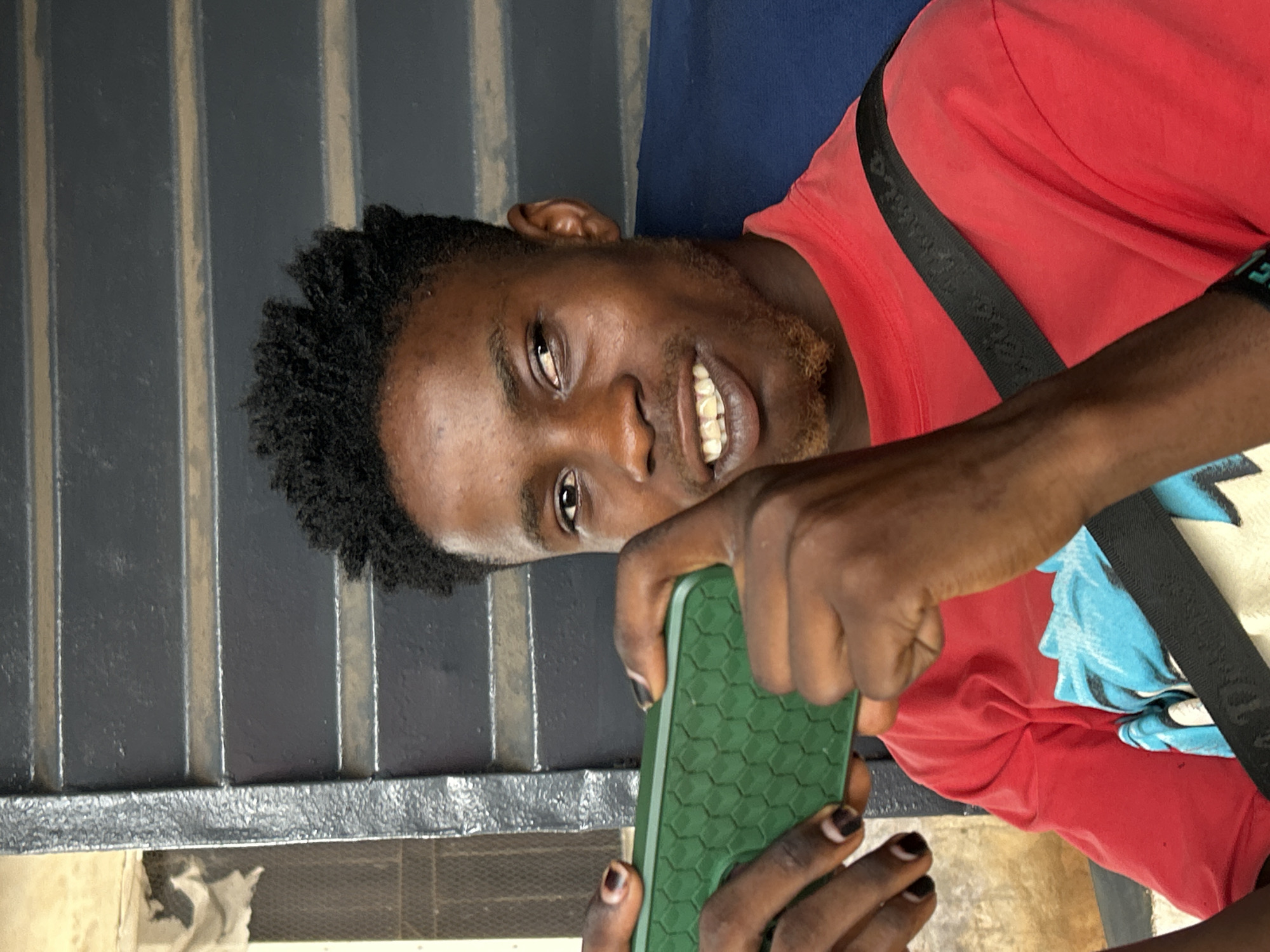 A Black man is holding a green cellular phone with a hexagonal pattern on its back, appearing to take a photograph. He has short, dark, textured hair and a beard with reddish tones. The man is wearing a red t-shirt with a black strap crossing his chest. His fingernails appear to be painted dark, possibly black. The background includes a dark gray corrugated metal surface and a patch of blue fabric. The location is not discernible from the image.