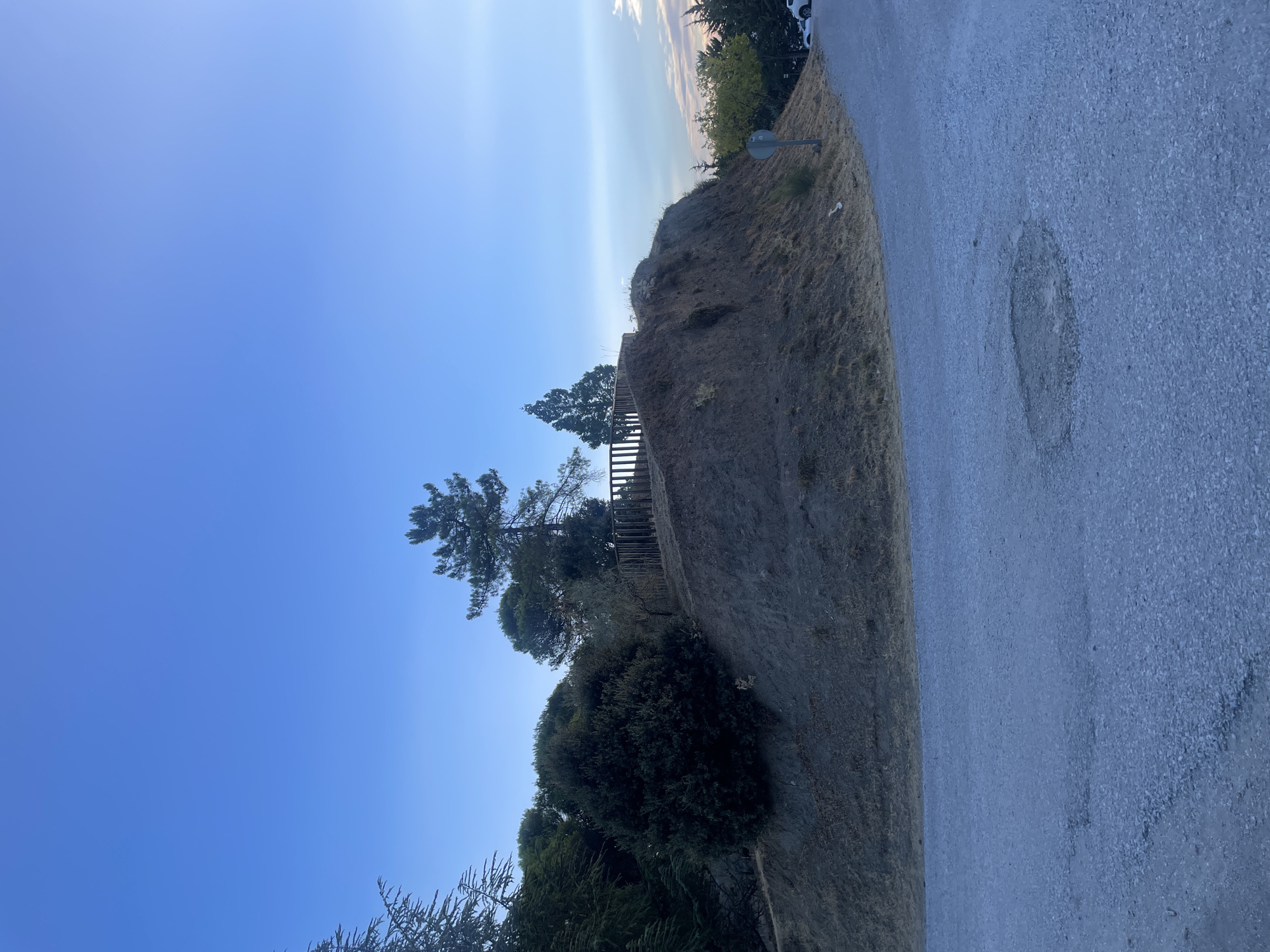 The image shows a slight incline with dry grass and a dirt path leading up to a small hill. At the top of the hill, there is a wooden fence and several trees. The sky is clear and blue, with a few wispy clouds on the horizon. In the foreground, there is a paved road with a patch of discolored asphalt. The scene appears to be in a rural or suburban area, possibly on the outskirts of the city of Víznar, Spain, given the arid landscape and vegetation. There are no people or animals visible in the image.