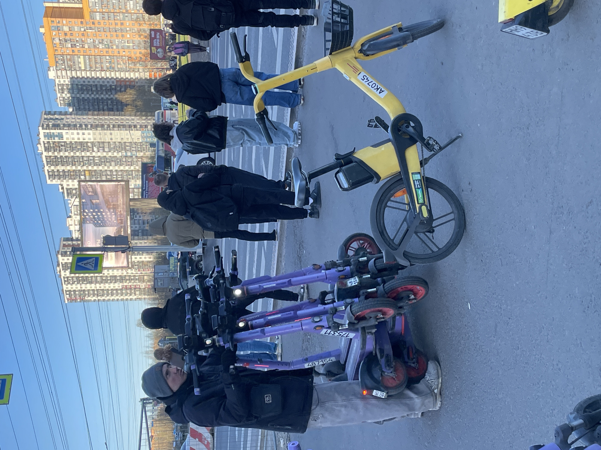 The image depicts a bustling urban street scene on a clear, sunny day. In the foreground, a person wearing a black jacket and a grey beanie stands near a cluster of several purple electric scooters. These scooters bear visible identification numbers such as "175311" and "487154," along with "UGO" and "YOU" branding. To the right of the person, a bright yellow rental bicycle is prominently parked, displaying "AK0745" on its frame and "BE 83" on its rear mudguard. A small screen near the bicycle's front wheel shows partial Cyrillic text, possibly "Право" (meaning "Right").
Several other individuals are visible in the middle ground and background, walking or standing on the street, which features white pedestrian crossing stripes. They are dressed in casual attire, including jackets and jeans.
The background is dominated by a bright blue sky and numerous tall residential buildings, varying in color from light to orange-brown. Power lines stretch across the upper part of the frame. Urban infrastructure such as a pedestrian crossing sign, a traffic light, and multiple billboards are present. Visible text on the billboards includes partially legible Cyrillic words like "РЫ" (likely part of "КВАРТИРЫ" - apartments), "ТАРТ", "АРОМА," and "СКИДКИ ВЫГОДНЫЕ..." (profitable discounts). The scene conveys an active, city environment.