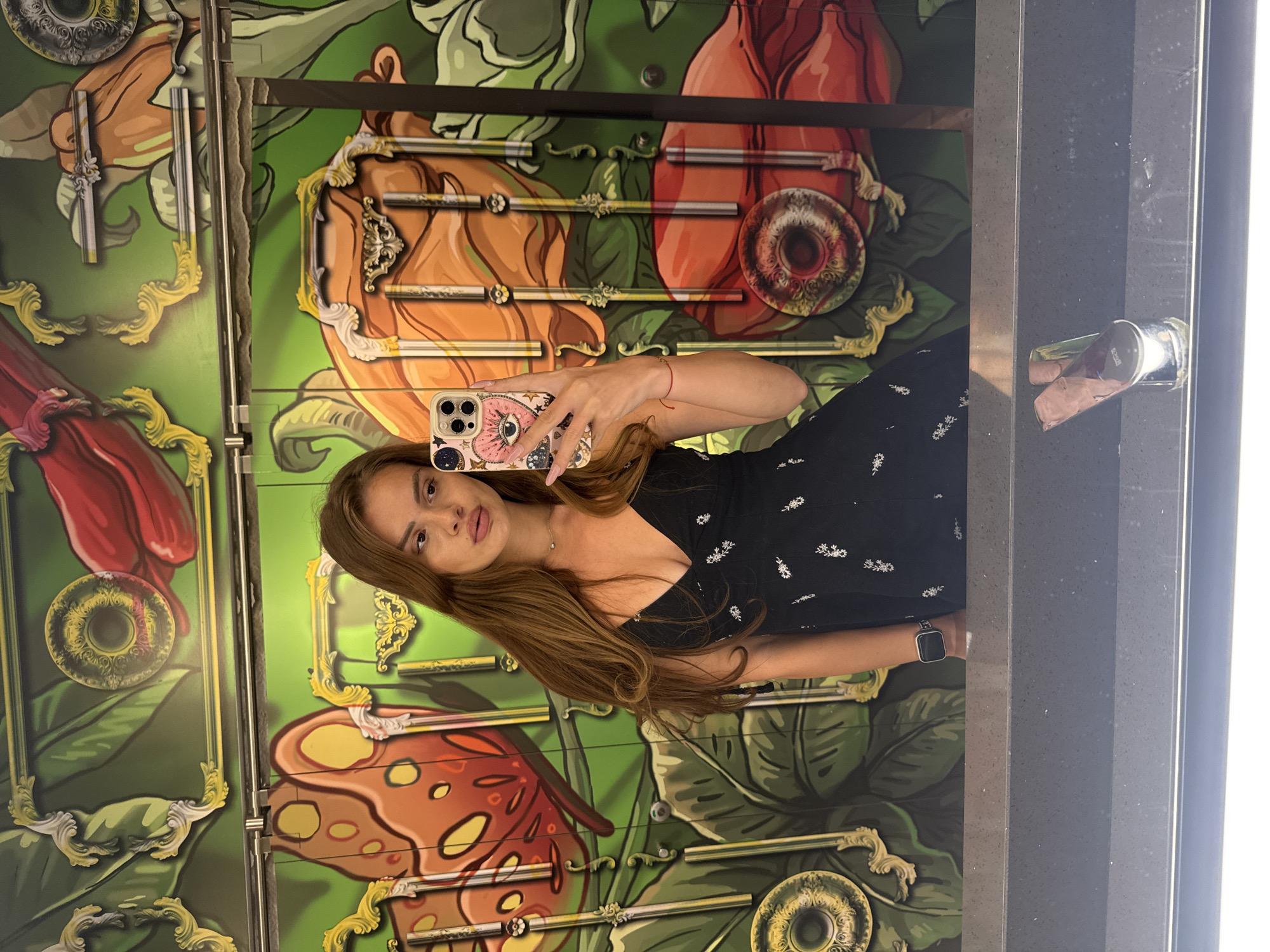 The media file is a vertically oriented, full-body, mirrored selfie taken by a young woman standing in front of a colorful mural. The woman has long, brown hair and is wearing a black dress with white floral patterns and a smartwatch on her left wrist. She is holding a pink smartphone with a decorative case featuring an eye motif and stars. Her right arm is extended, capturing the reflection.
The mural behind her is vibrant and features stylized botanical elements with shades of green, orange, and red. There are also decorative gold accents and ornate frames within the mural. The overall impression of the mural is whimsical and somewhat surreal.
The scene appears to be indoors, possibly in a public space or an establishment with this unique mural artwork. The lighting suggests it could be daytime. The woman's expression is neutral, and she is looking directly at the camera. The reflection of a cylindrical object, possibly a can, is visible in a darker, textured surface to the right of the mural, which could be part of the surrounding architecture or furniture. There is no visible text in the image. The context provided indicates the location as Plovdiv, Bulgaria.